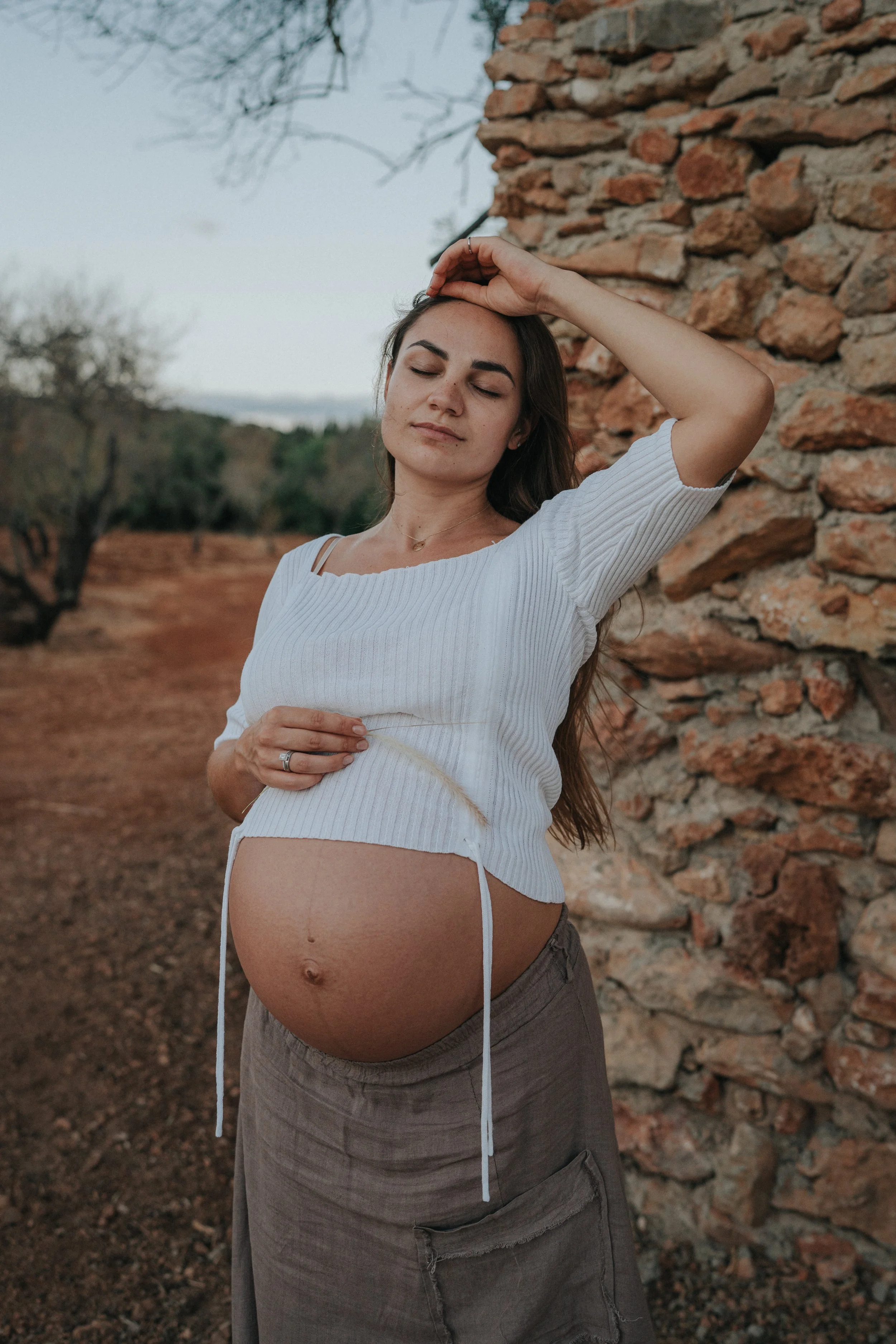 A pregnant woman with long brown hair, wearing a white cropped top and gray cargo pants, stands outdoors near a stone wall with her eyes closed and her hand resting on her forehead. The setting appears to be a rural area with trees and a dirt path.