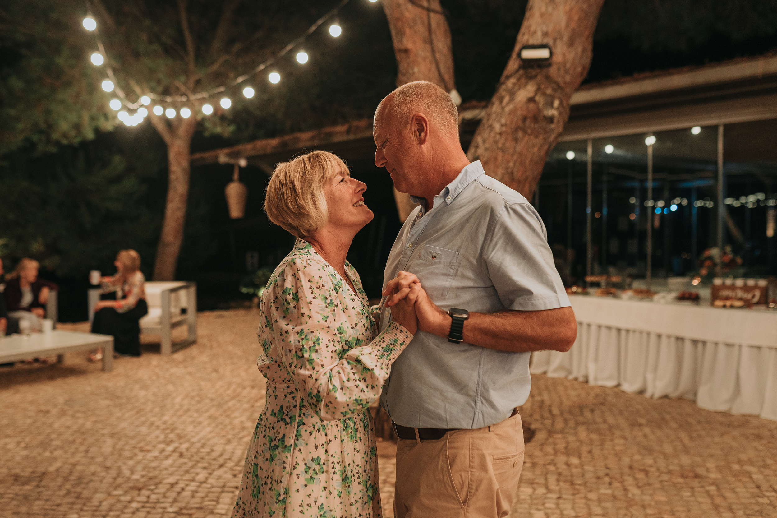 A couple is dancing closely at an outdoor evening event. The surroundings are decorated with string lights and lanterns, with other people seated and enjoying the occasion in the background.