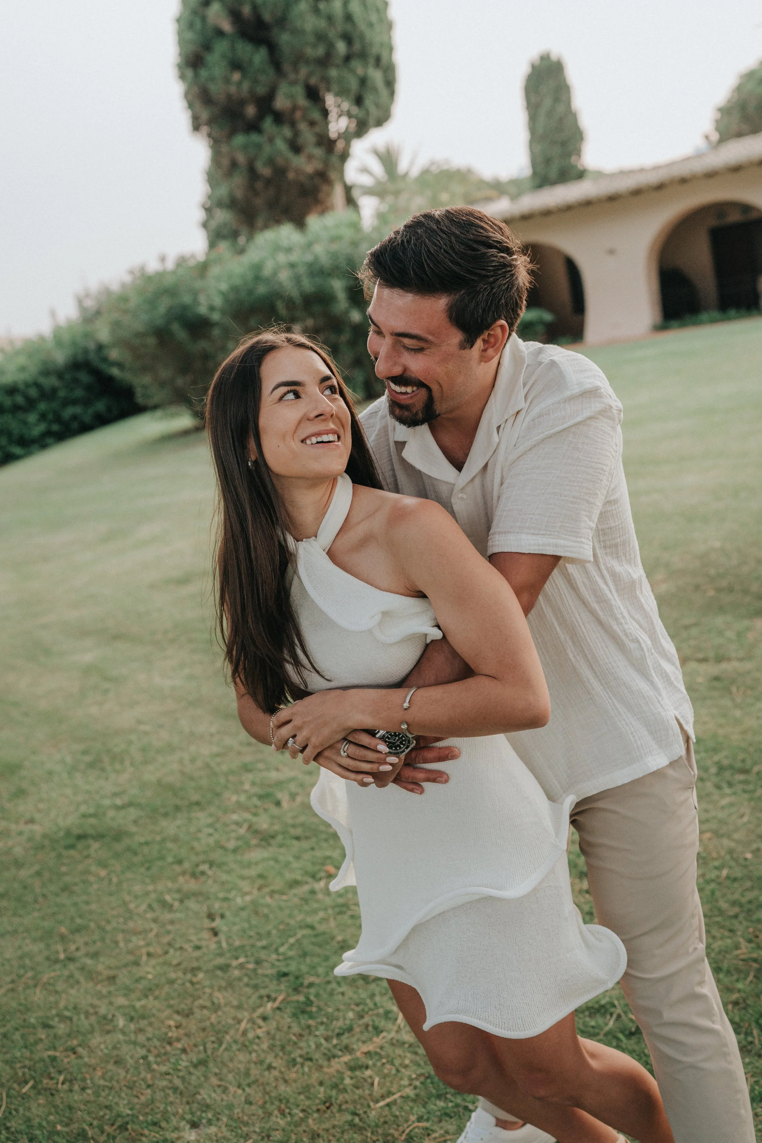 A man and a woman are dancing and smiling on a grassy lawn, with the woman in a white dress and the man in a beige shirt and pants, with trees and a building in the background.