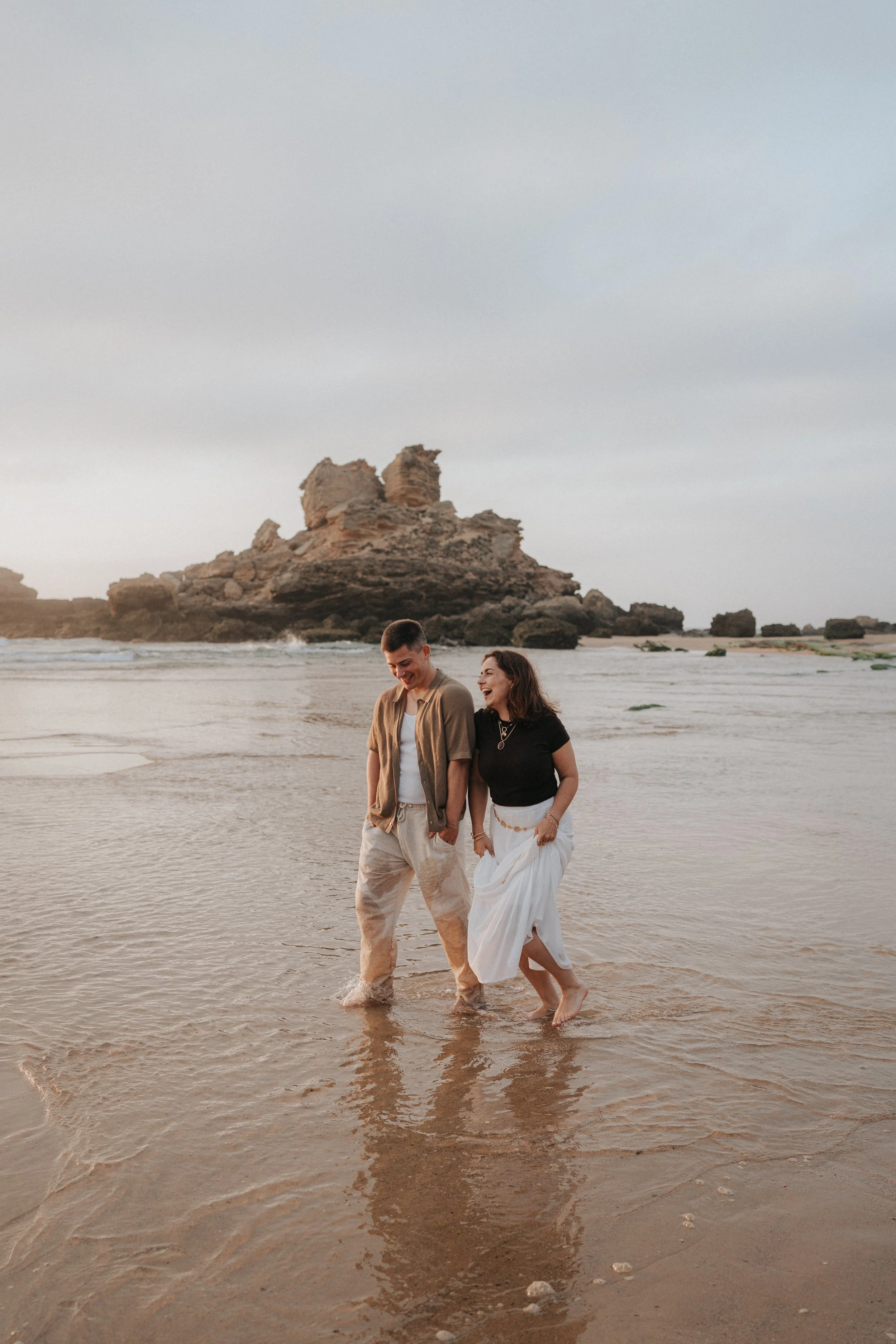 A couple walking barefoot on the beach near the water, smiling and looking at each other, with rocky formations in the background under a cloudy sky.