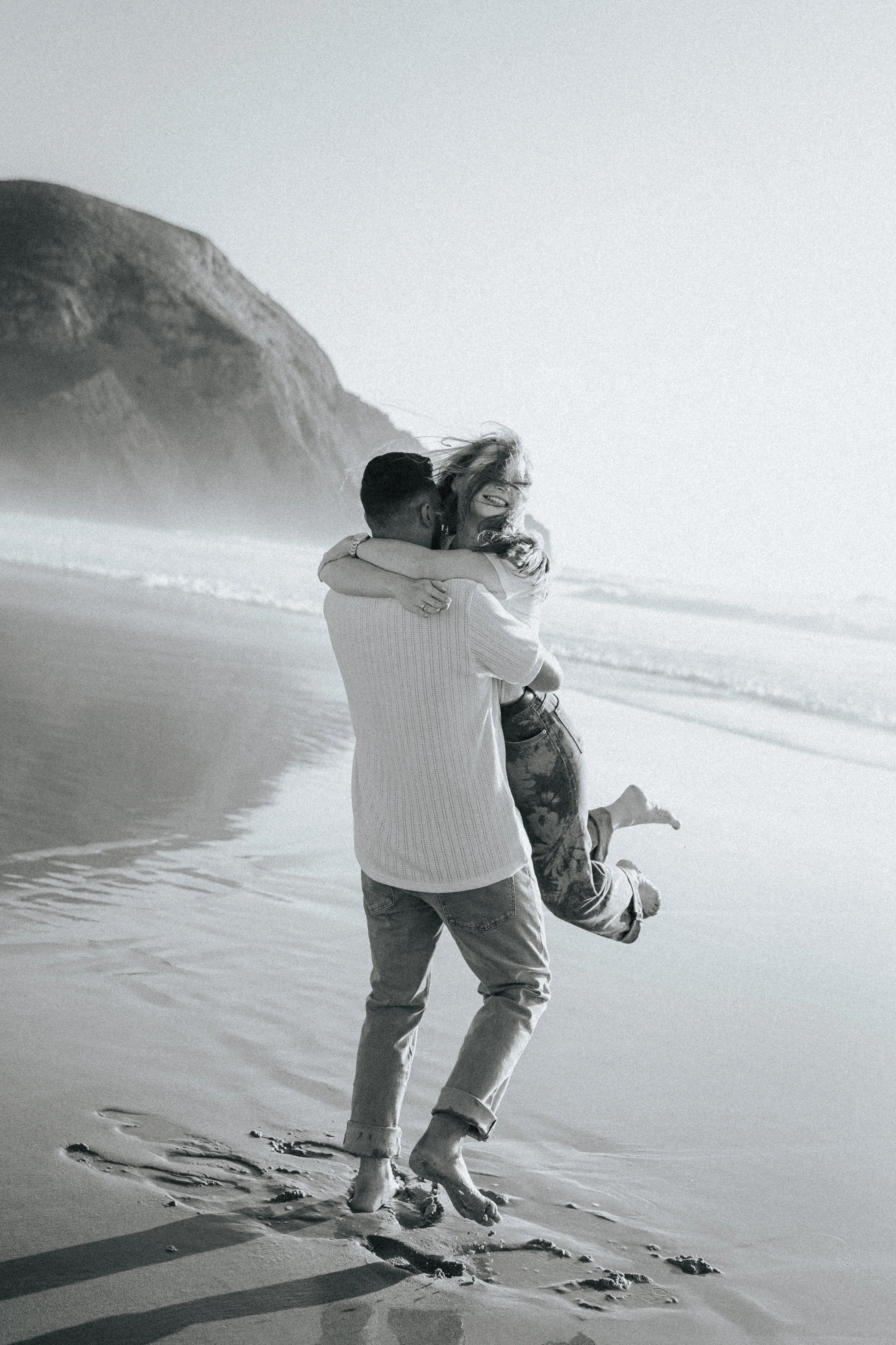 A couple embracing on the beach, with the man holding the woman in his arms, as they laugh and kiss. The woman has long hair and is wearing a patterned outfit, and the man is wearing a light-colored sweater and jeans. They are walking along the shore