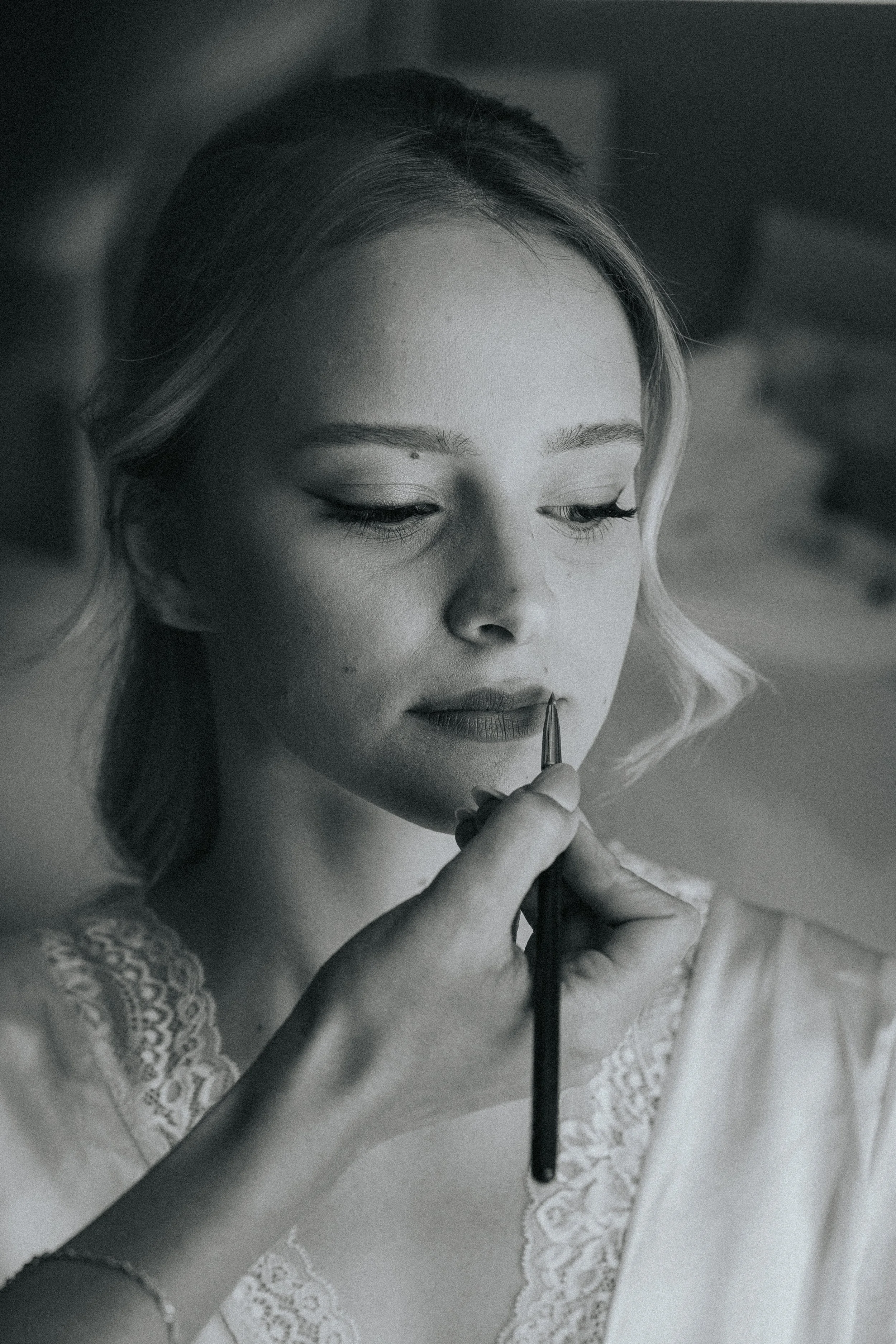 A woman with light skin and lace clothing is holding a makeup brush close to her face, looking down with a soft expression.