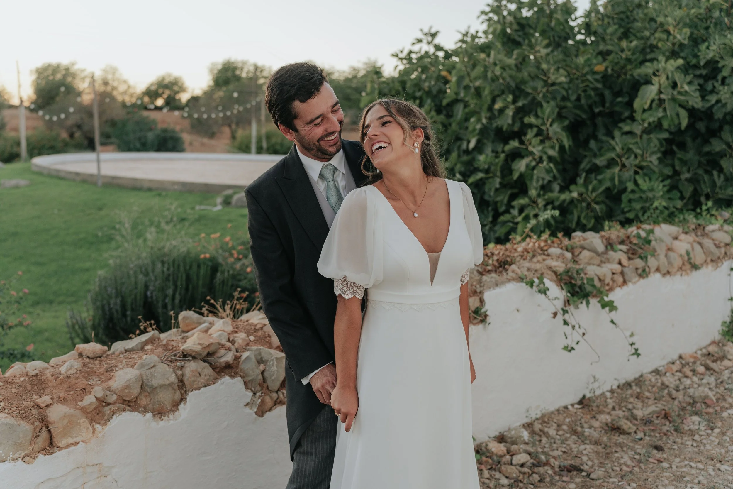 A newlywed couple smiling and enjoying their wedding day outdoors with greenery and string lights in the background.