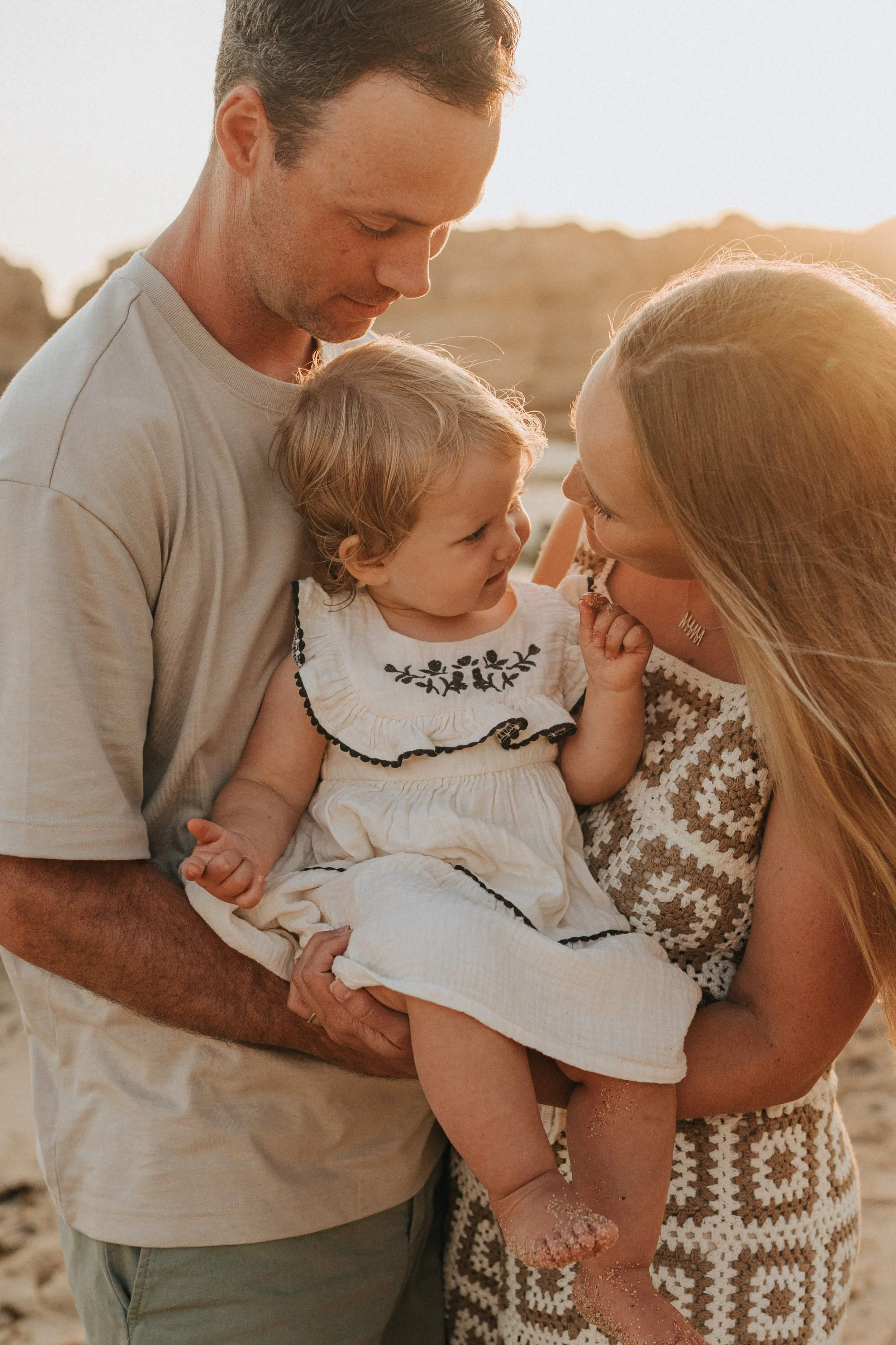 A family of three, a man, a woman, and a young girl, sharing a tender moment outdoors at sunset, with a blurred background of rocks or hills.