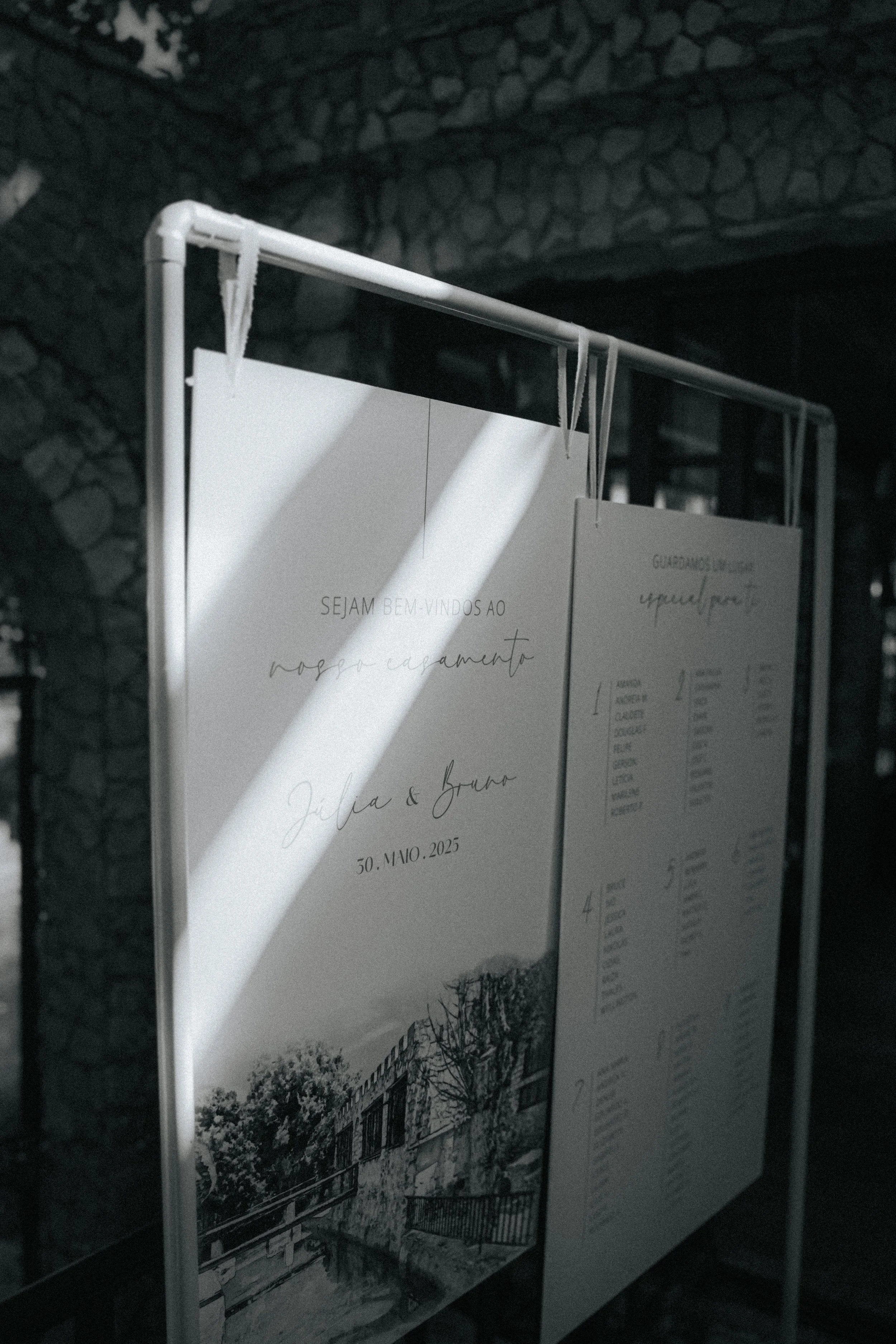 A wedding invitation poster hangs from a white metal stand, with a shadow cast onto it. The poster includes elegant script with the names Julia and Bruno, along with the date 30th of November, 2025.