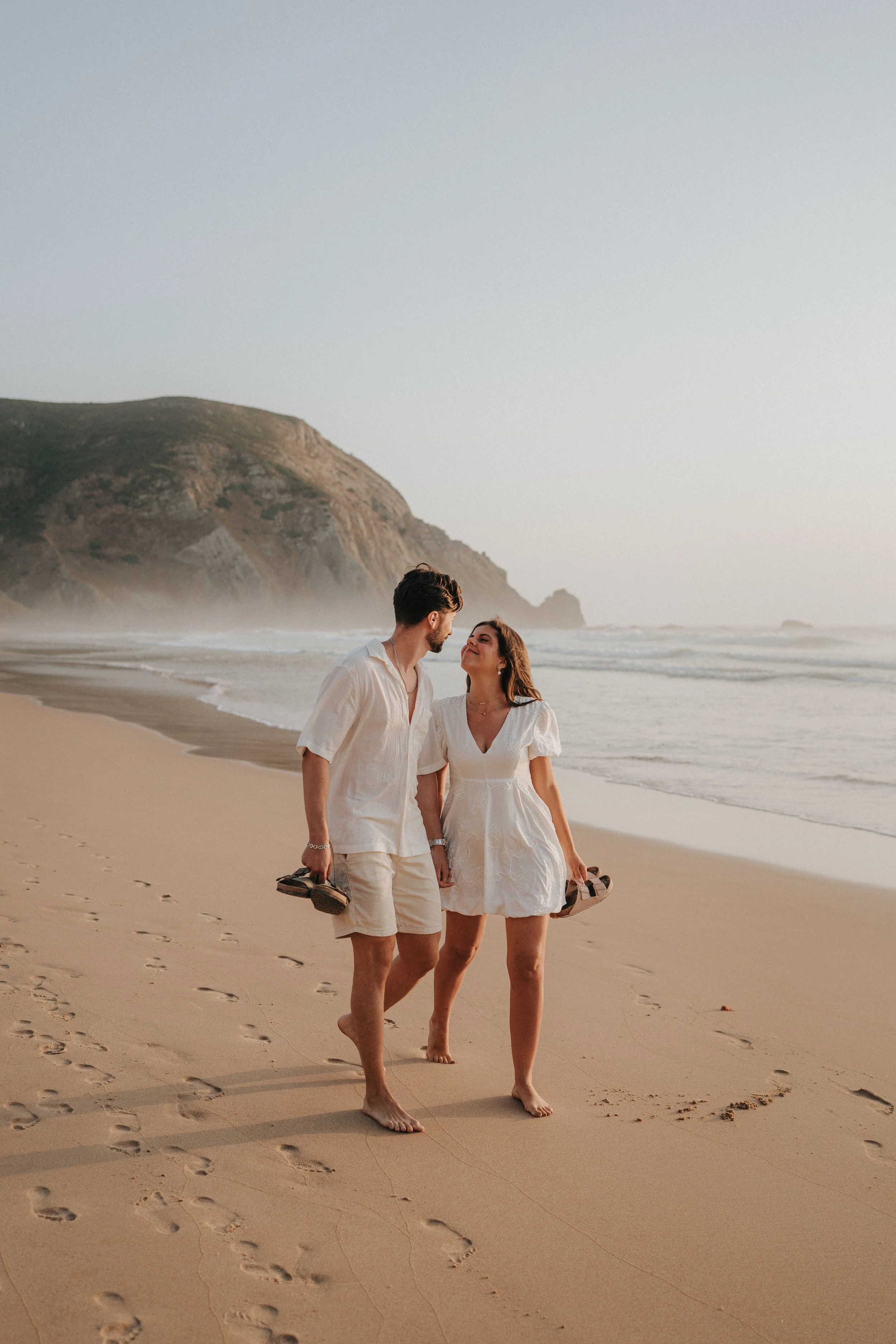 A couple walking on the beach holding hands, with sandals in hand, during sunset with cliffs in the background.