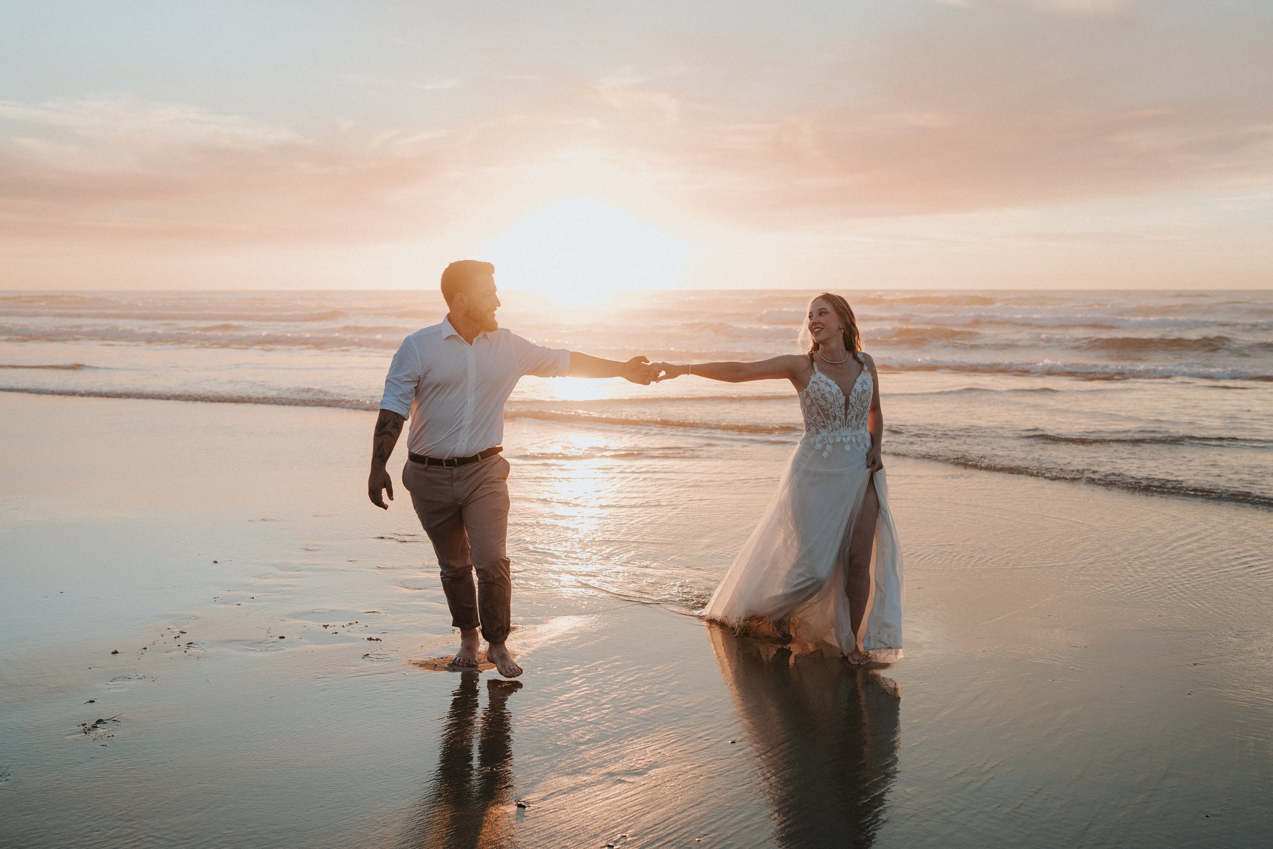 A couple holding hands and walking on the beach during sunset, with the ocean and sky in the background.