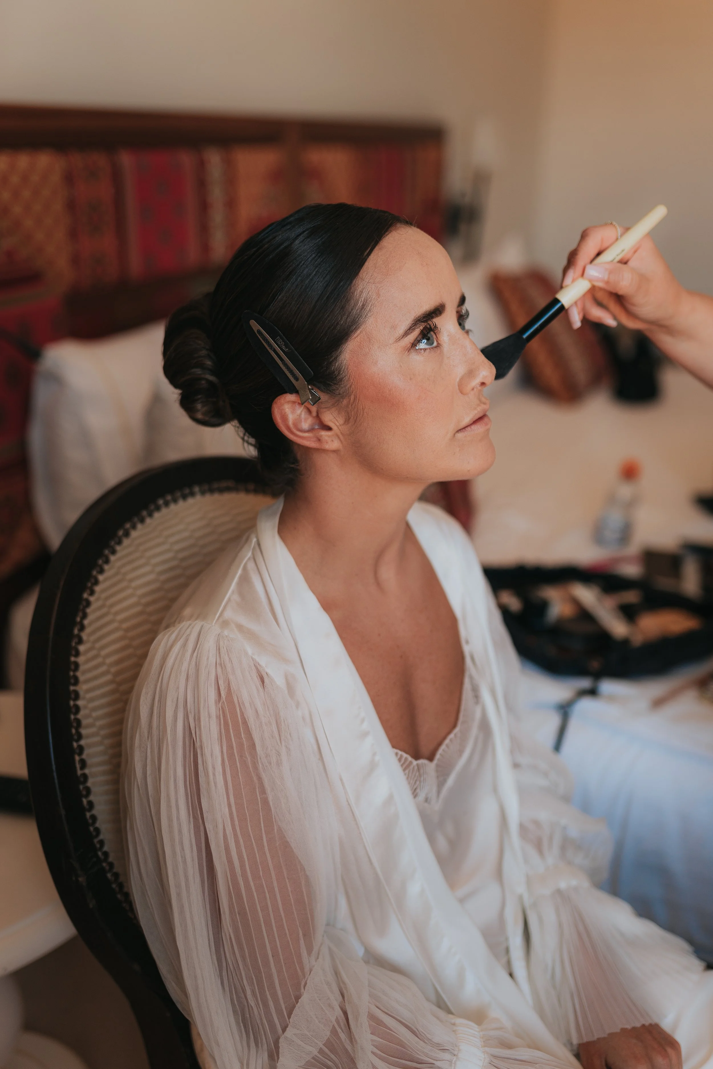 A woman with dark hair styled in a braid, wearing a white silky robe, is having makeup applied with a brush. She is sitting in a room with a patterned headboard and pillows in the background.