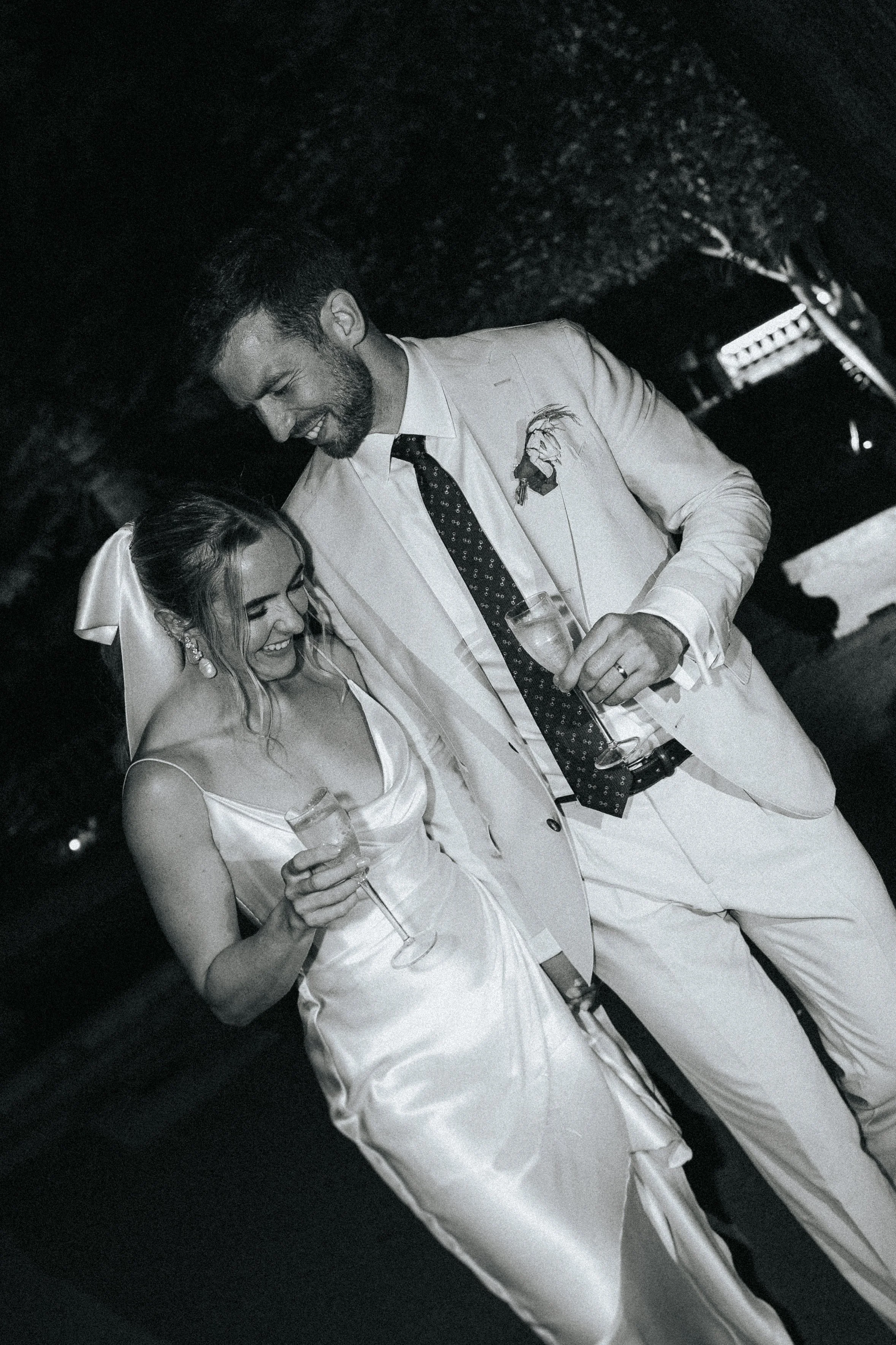 A man and woman smiling and looking down at each other at a formal event, with the man holding a drink and wearing a suit with a flower on the lapel, and the woman holding a glass, in black and white.