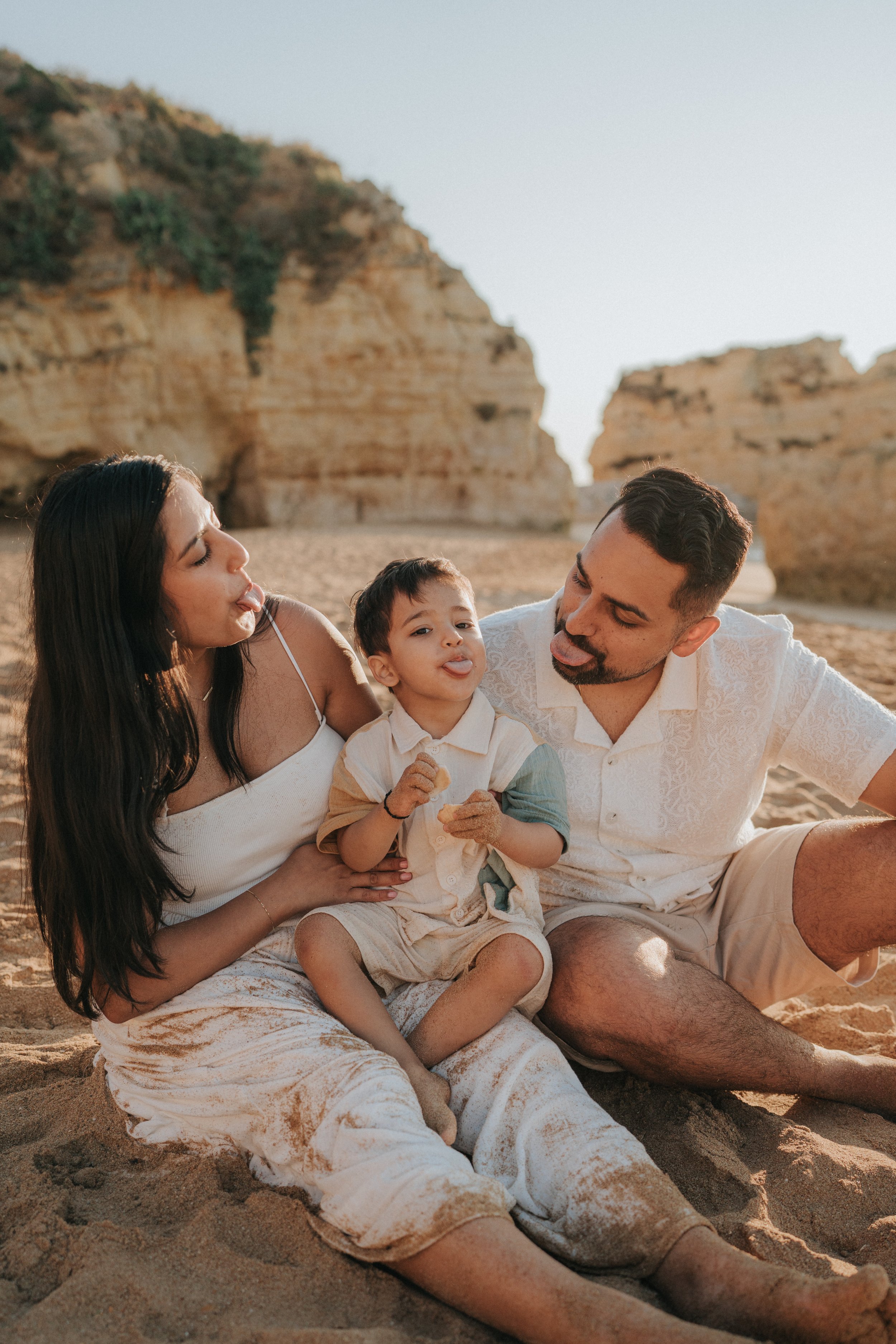 A family of three, a woman, a man, and a young boy, sitting on the sand at the beach with sandstone cliffs in the background. They are making playful faces, with the boy and woman sticking out their tongues.