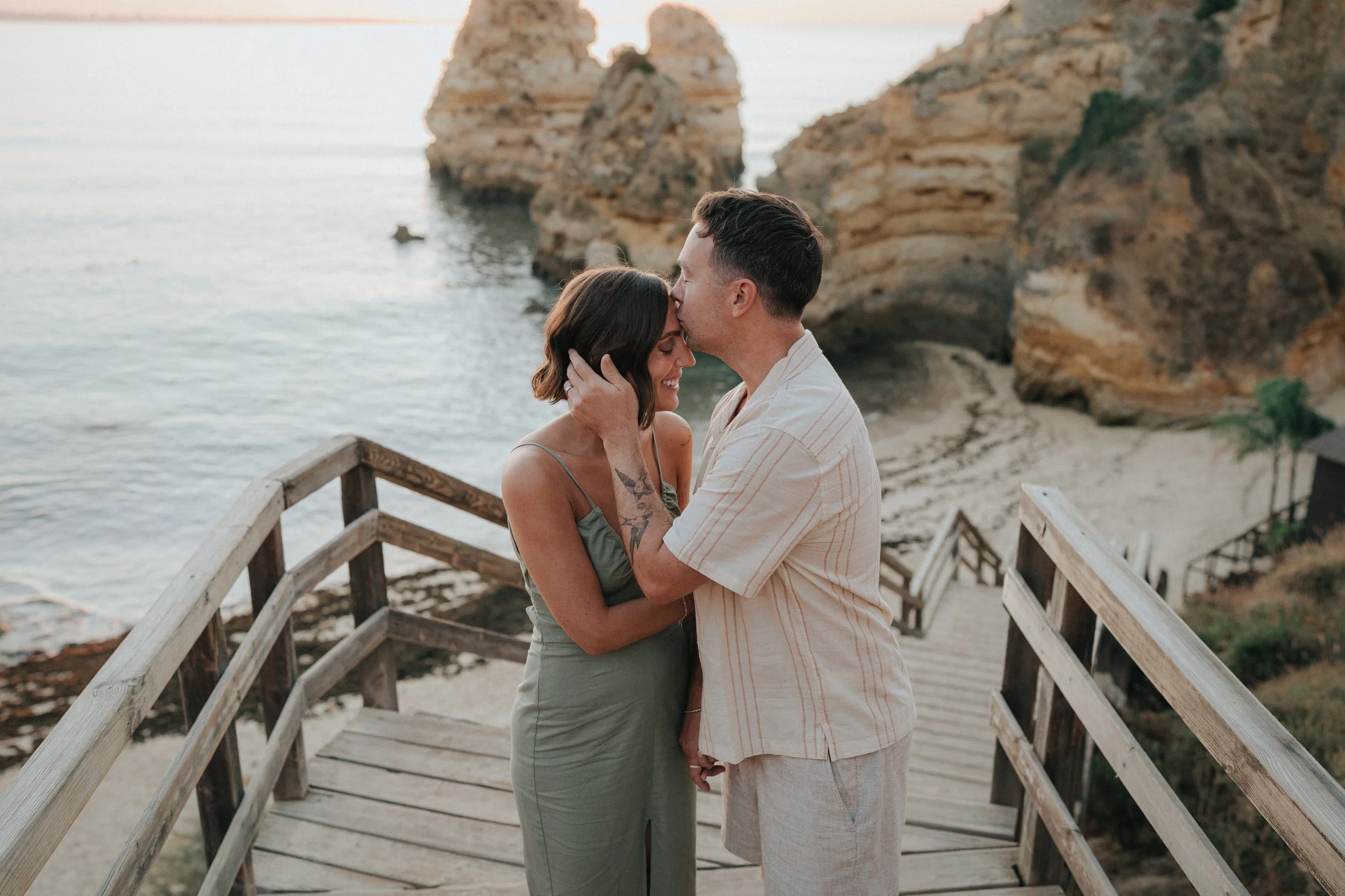 A couple standing on a wooden boardwalk by the beach, holding hands, with the man kissing the woman's forehead, overlooking rocky cliffs and the ocean at sunset.