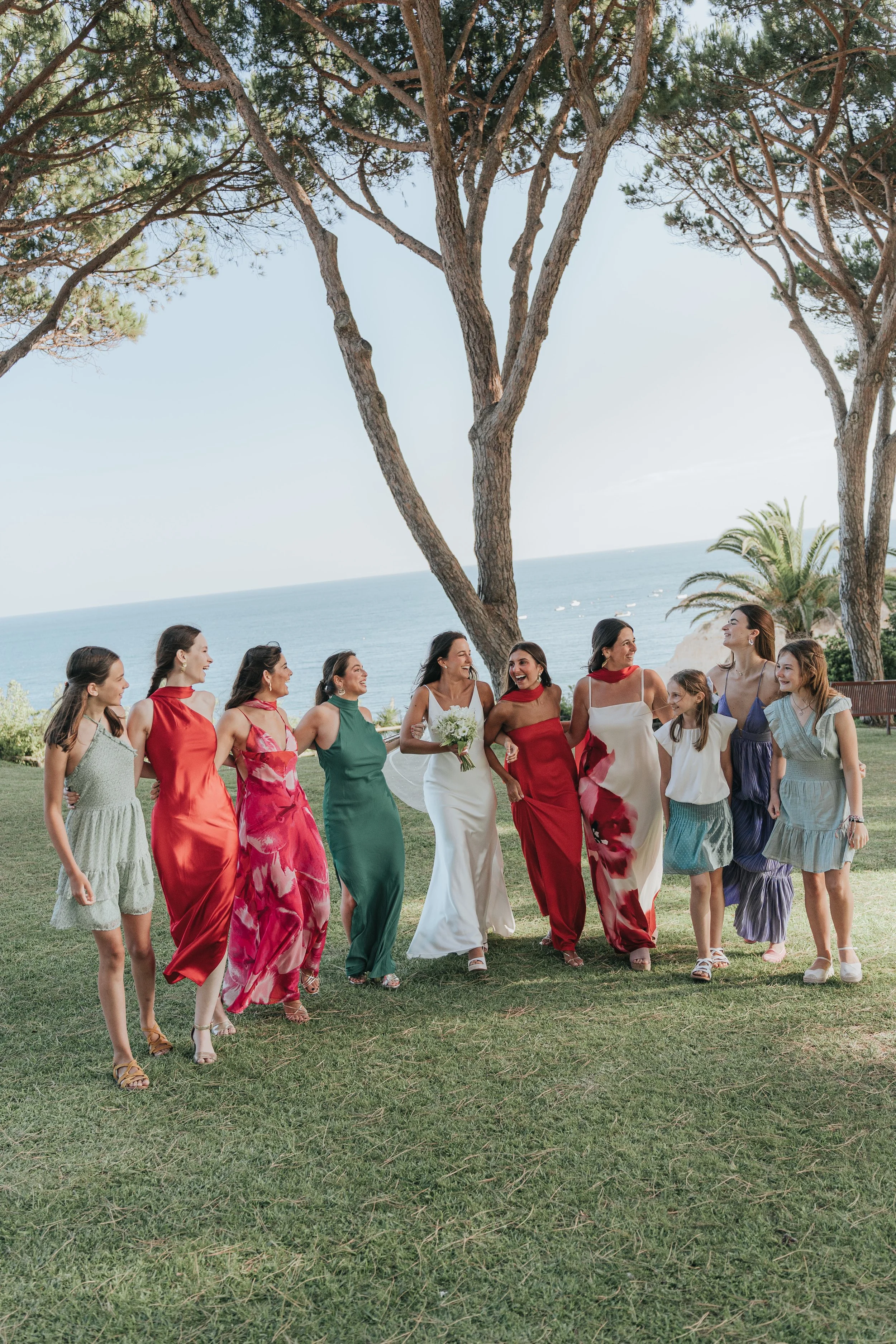 A group of women and children walking together outdoors near the ocean, with trees and palm trees in the background, during sunny weather.
