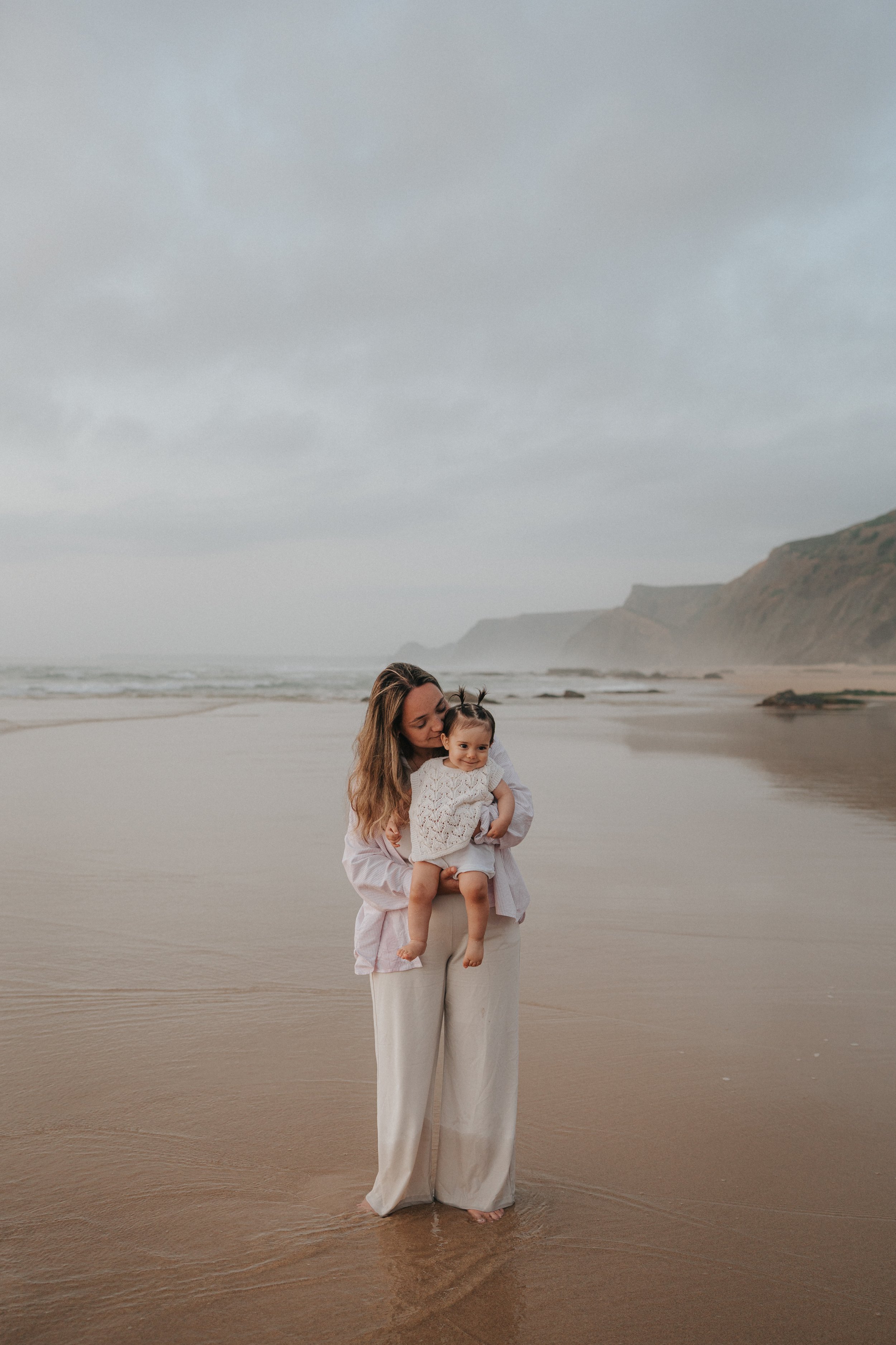 A woman holding a smiling child at the beach with cliffs in the background and overcast skies.