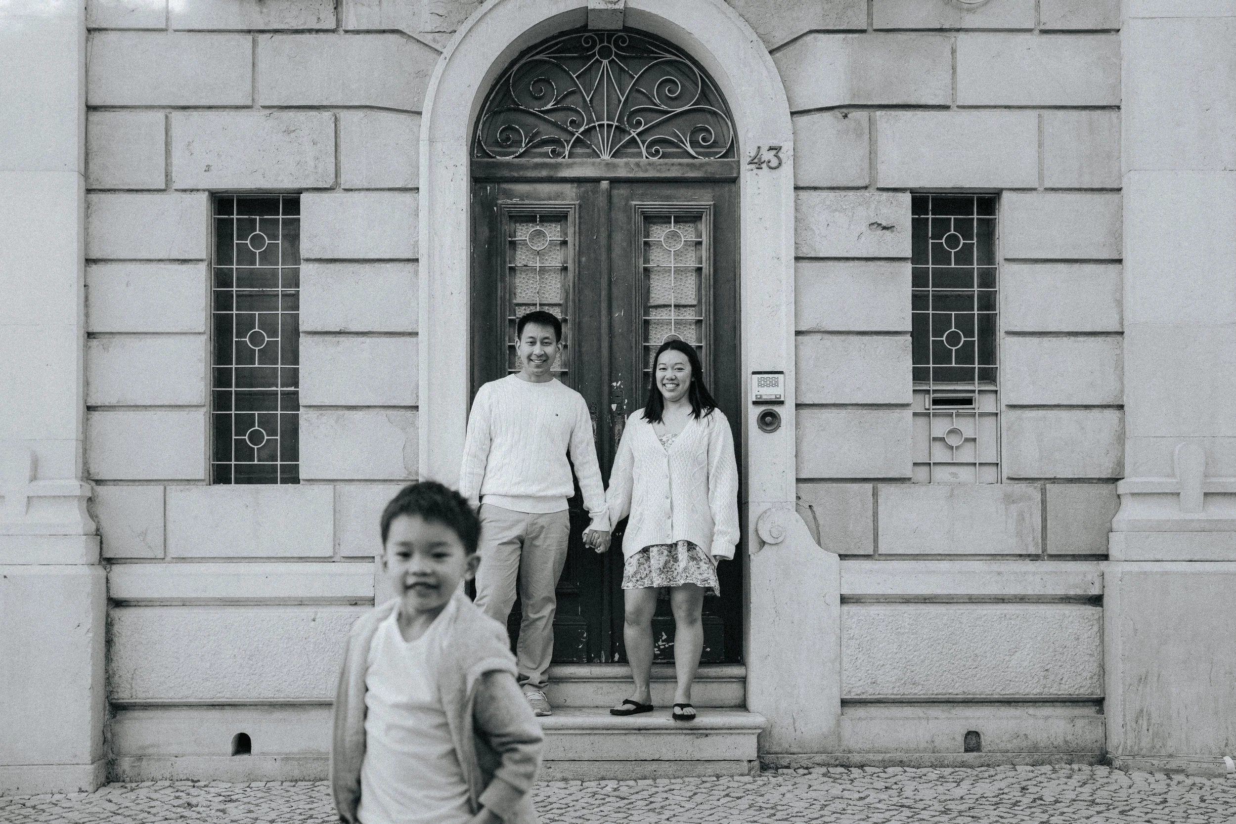 A family of four with two adults and two children standing on the steps of a building with a stone facade and decorative windows. The adults are holding hands, and the young boy is in the foreground, smiling and running toward the camera. The buildin