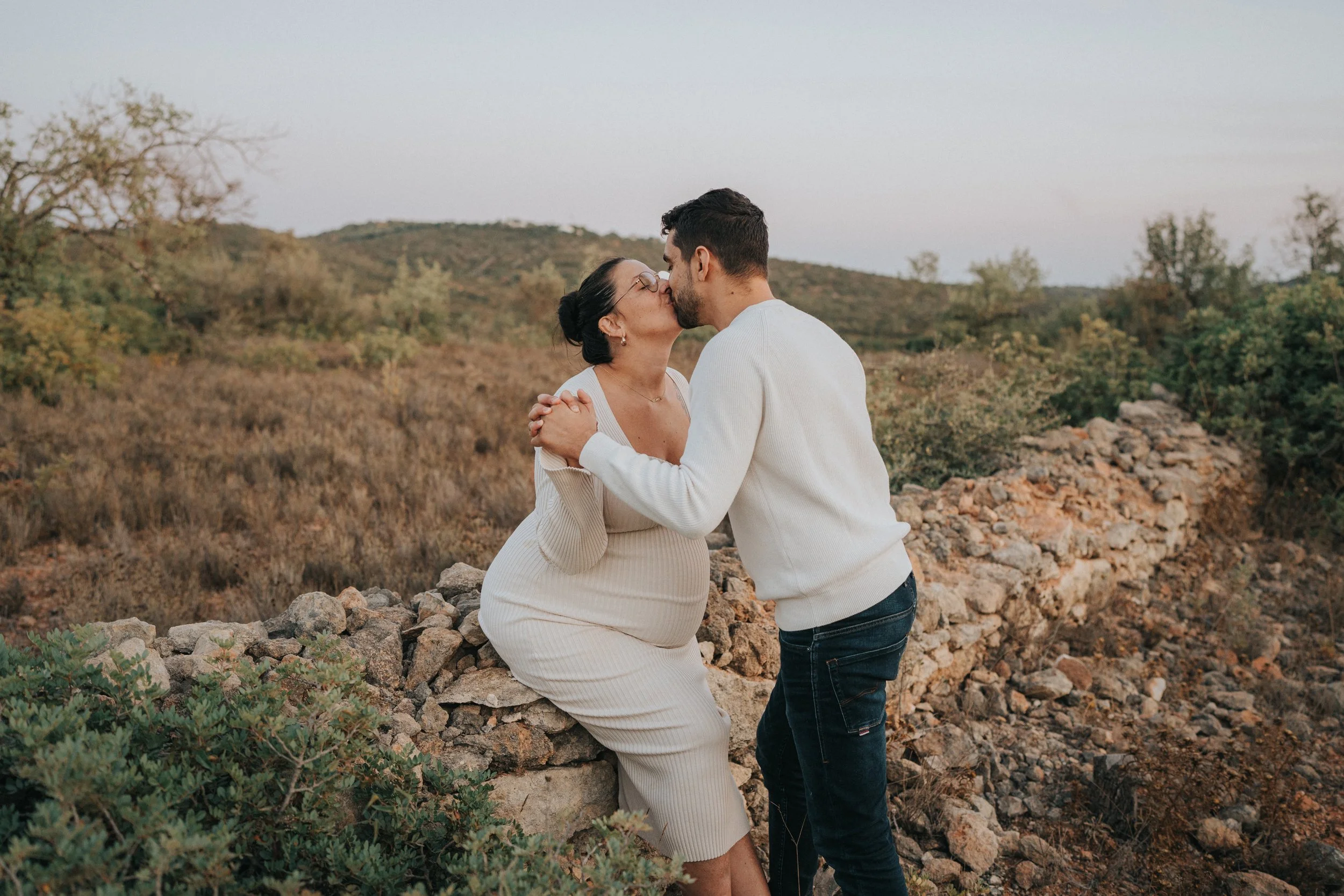 A man and woman share a kiss outdoors on a rocky area with hills and sparse trees in the background.