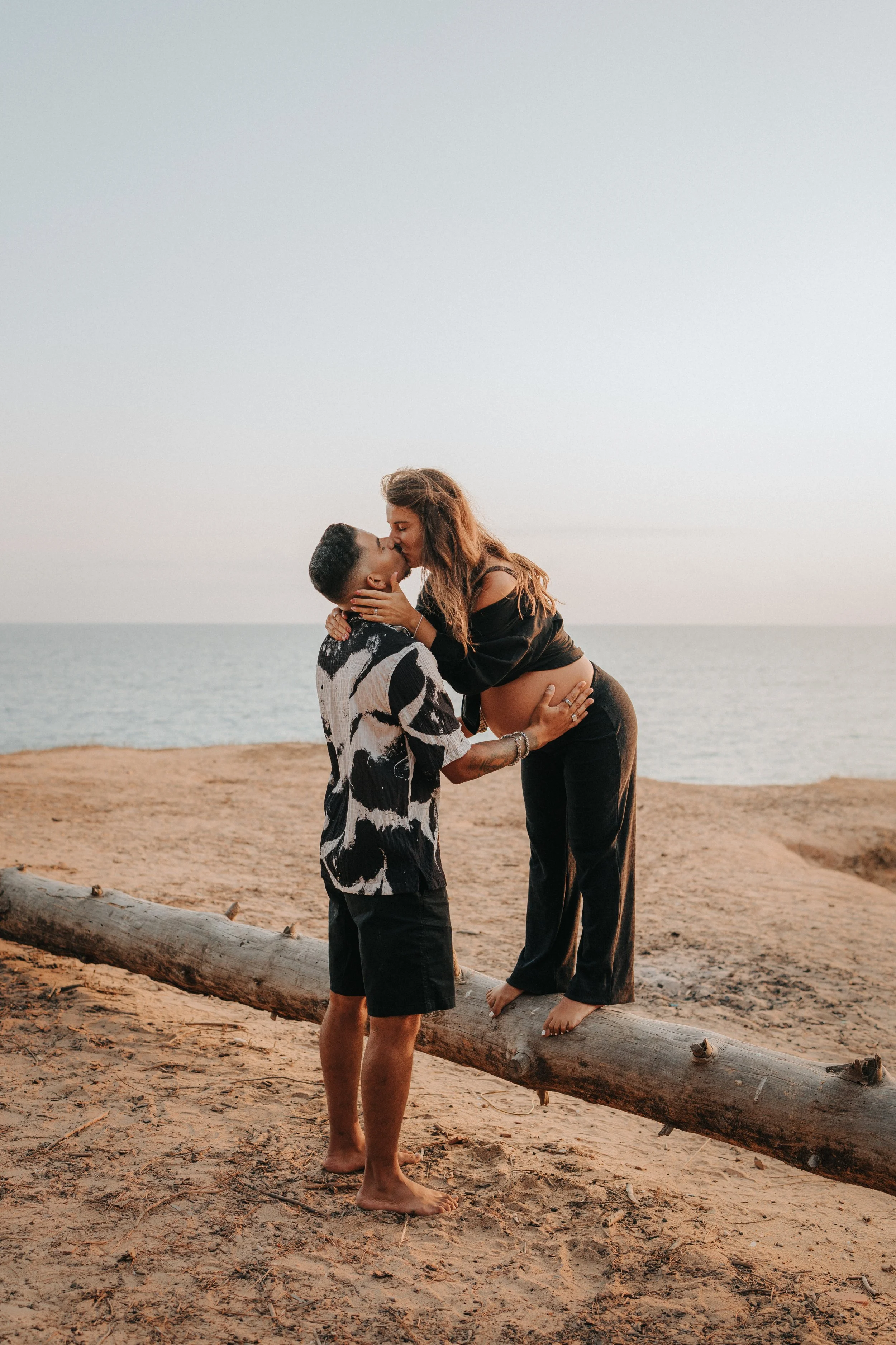 A couple sharing a kiss by the ocean, standing on a drift log on a sandy beach, during sunset.