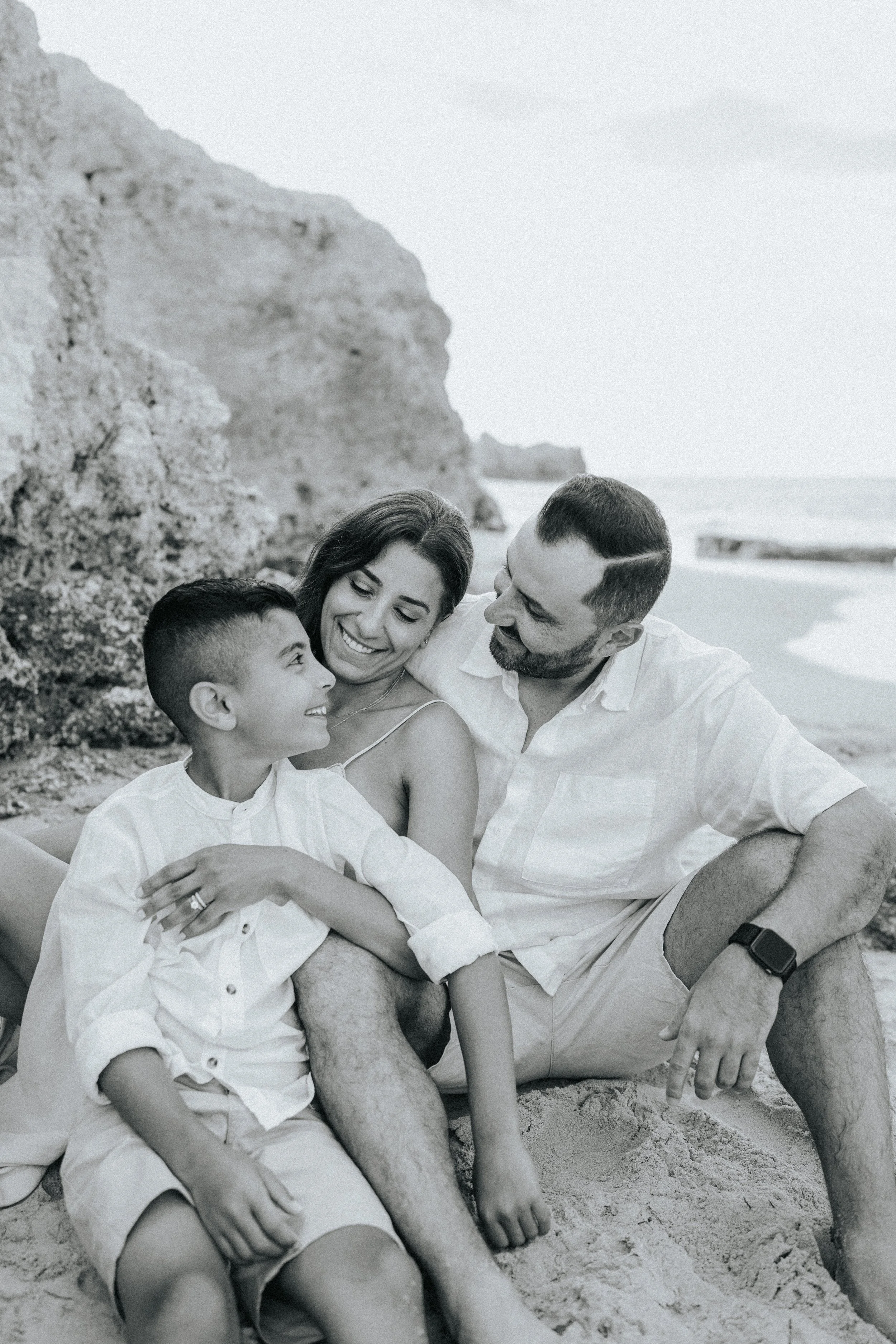 A happy family of four, including a woman, a man, and two young boys, sitting on the beach with rocks and ocean in the background. They are smiling and enjoying quality time together.