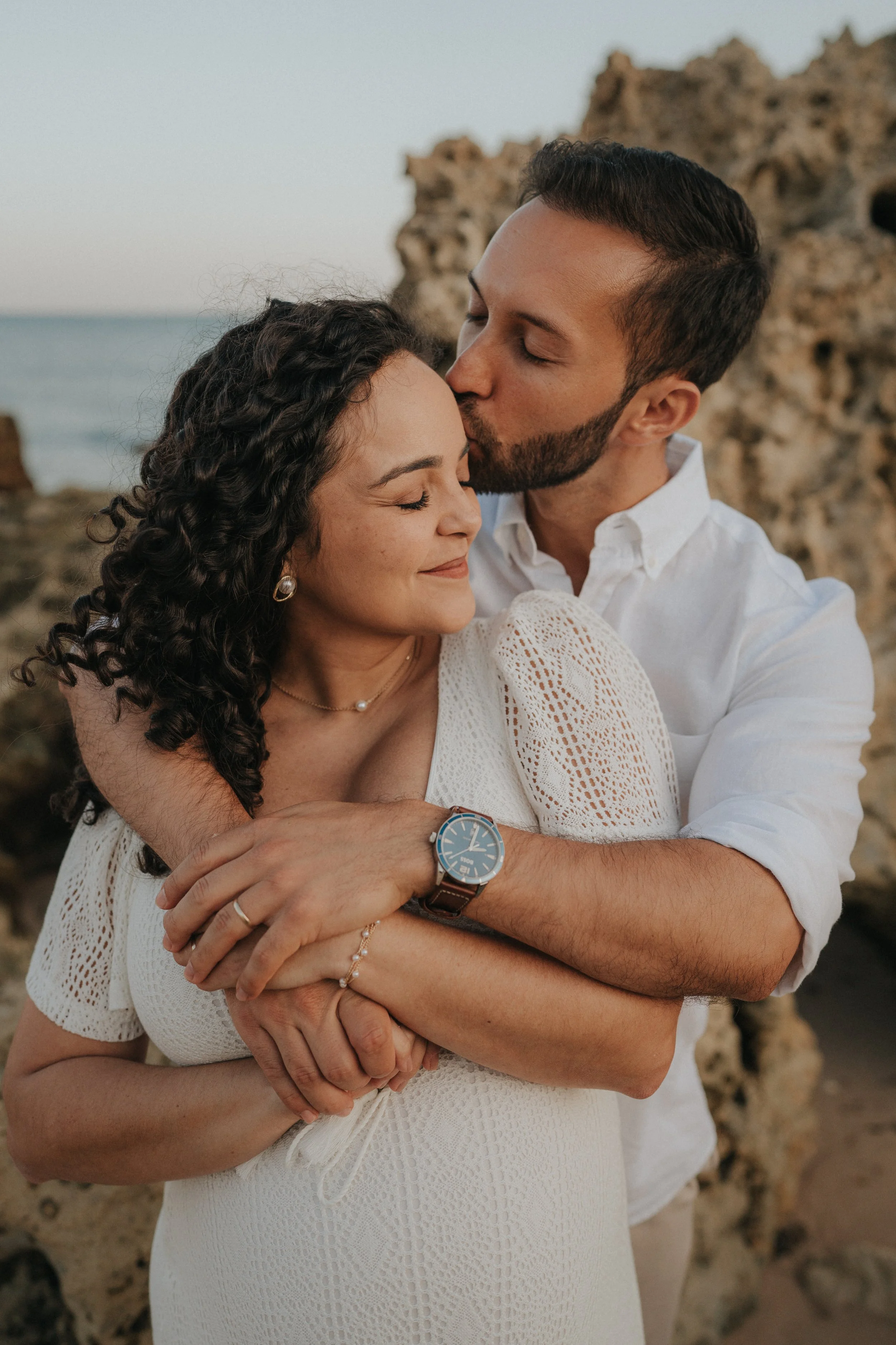 A couple embraces on a beach, the man kissing the woman on the forehead while she closes her eyes, both smiling gently. A rocky formation and the ocean are in the background.