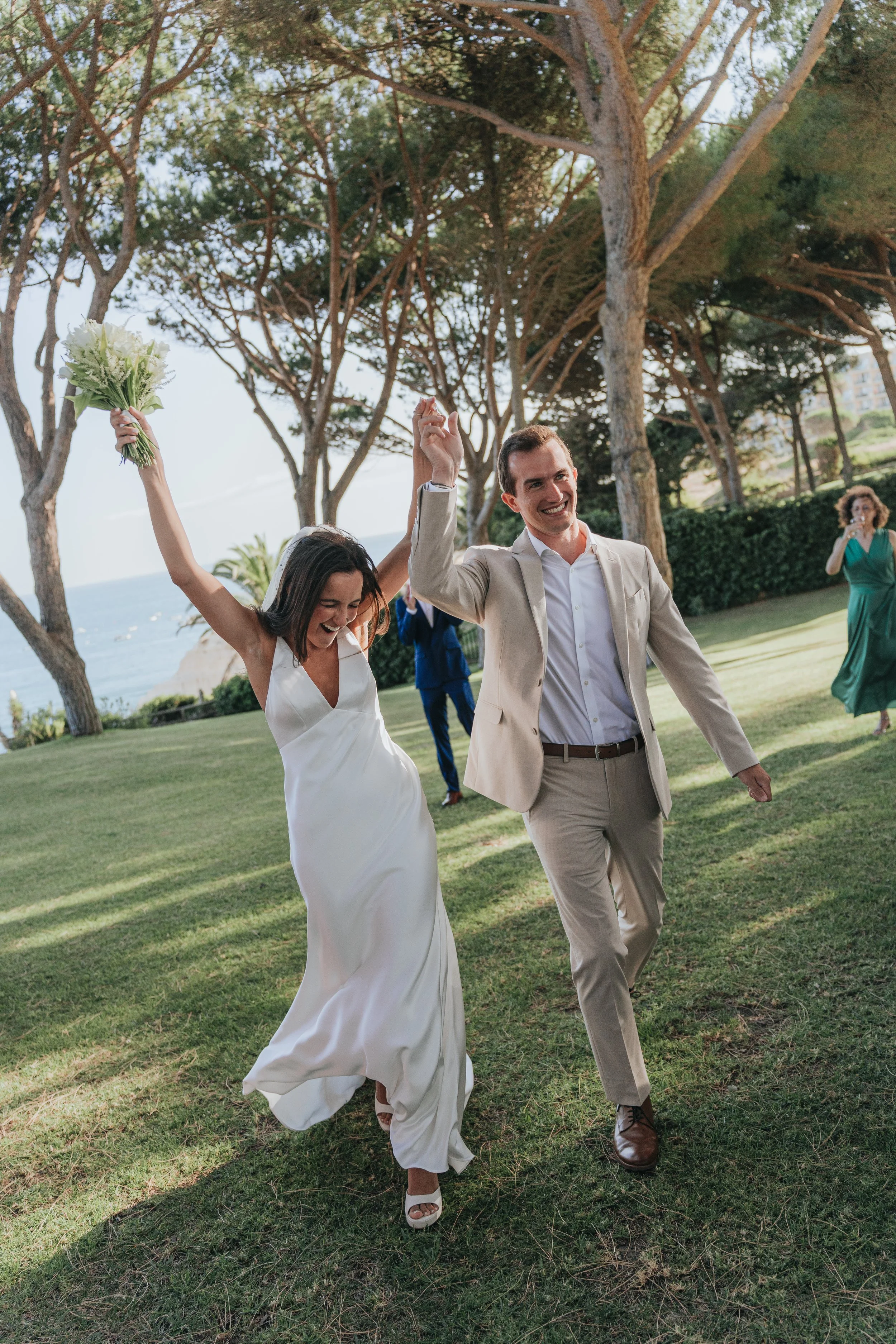 A woman in a white wedding dress and a man in a beige suit are celebrating outdoors under trees, holding hands up, with the woman holding a bouquet of flowers.