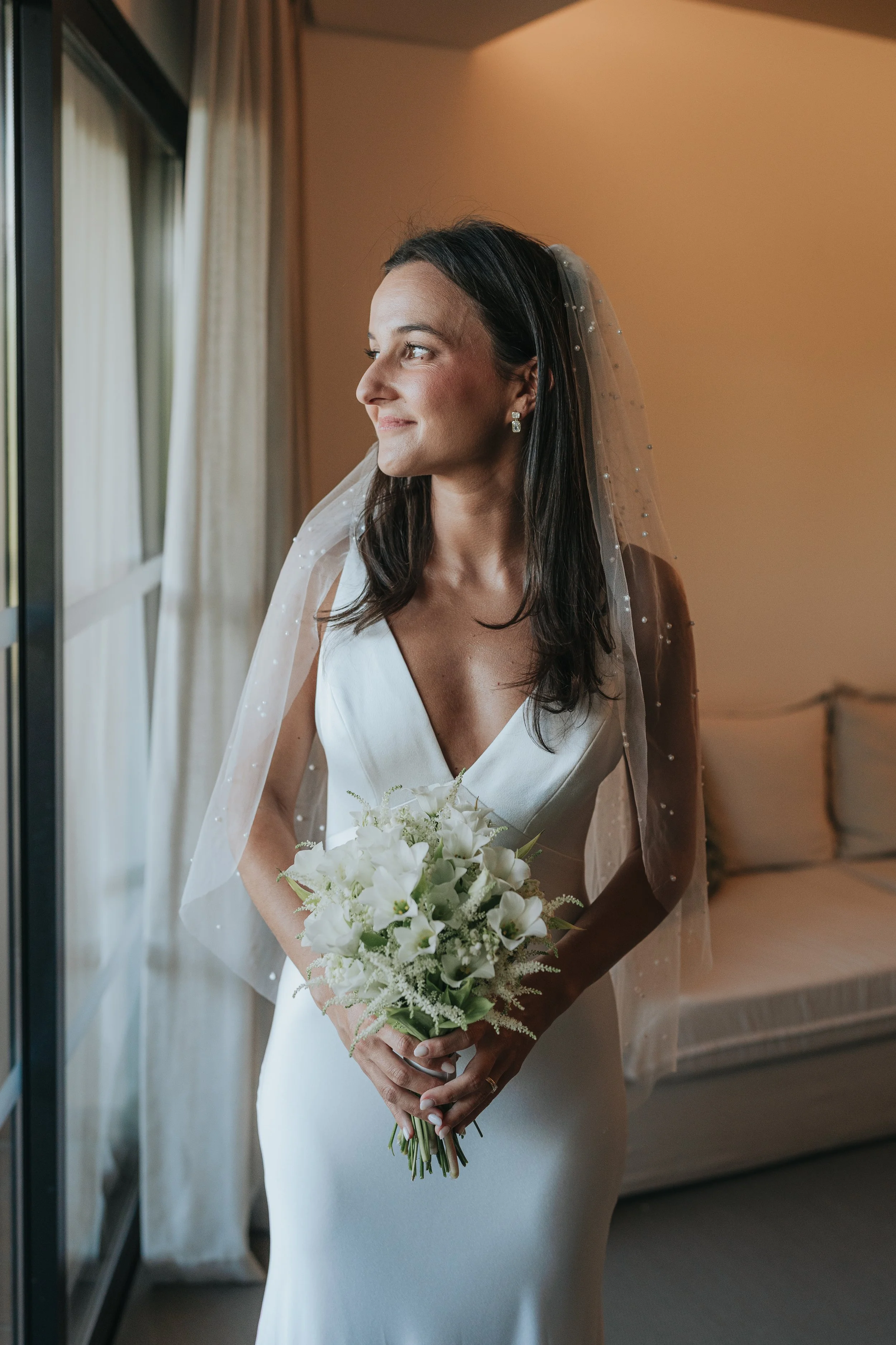 A bride with dark hair in a white wedding dress holding a bouquet of white flowers, standing indoors near a window with curtains, looking to her right with a gentle smile.