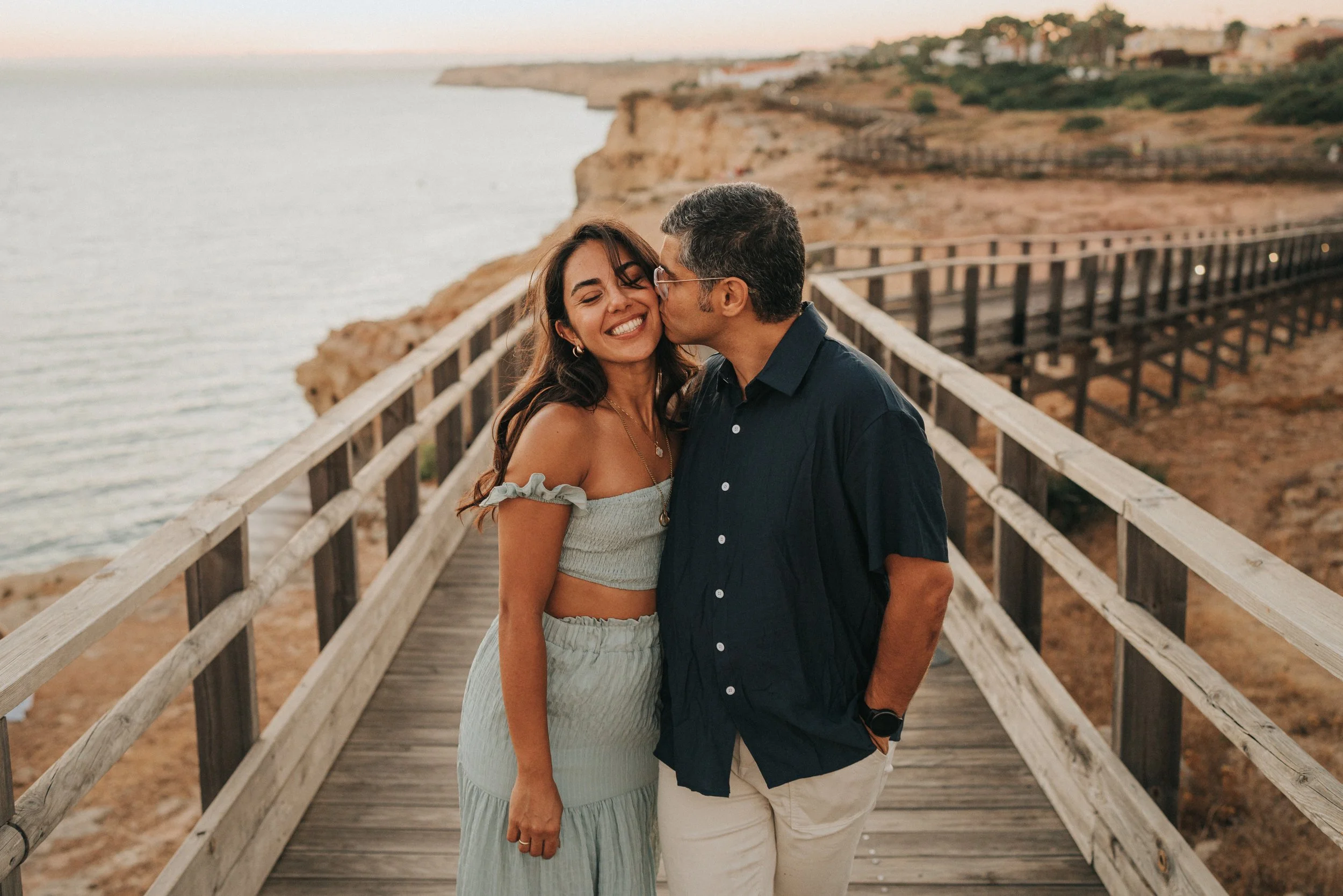 A smiling woman and a man share a kiss on the cheek on a wooden boardwalk overlooking the ocean at sunset.