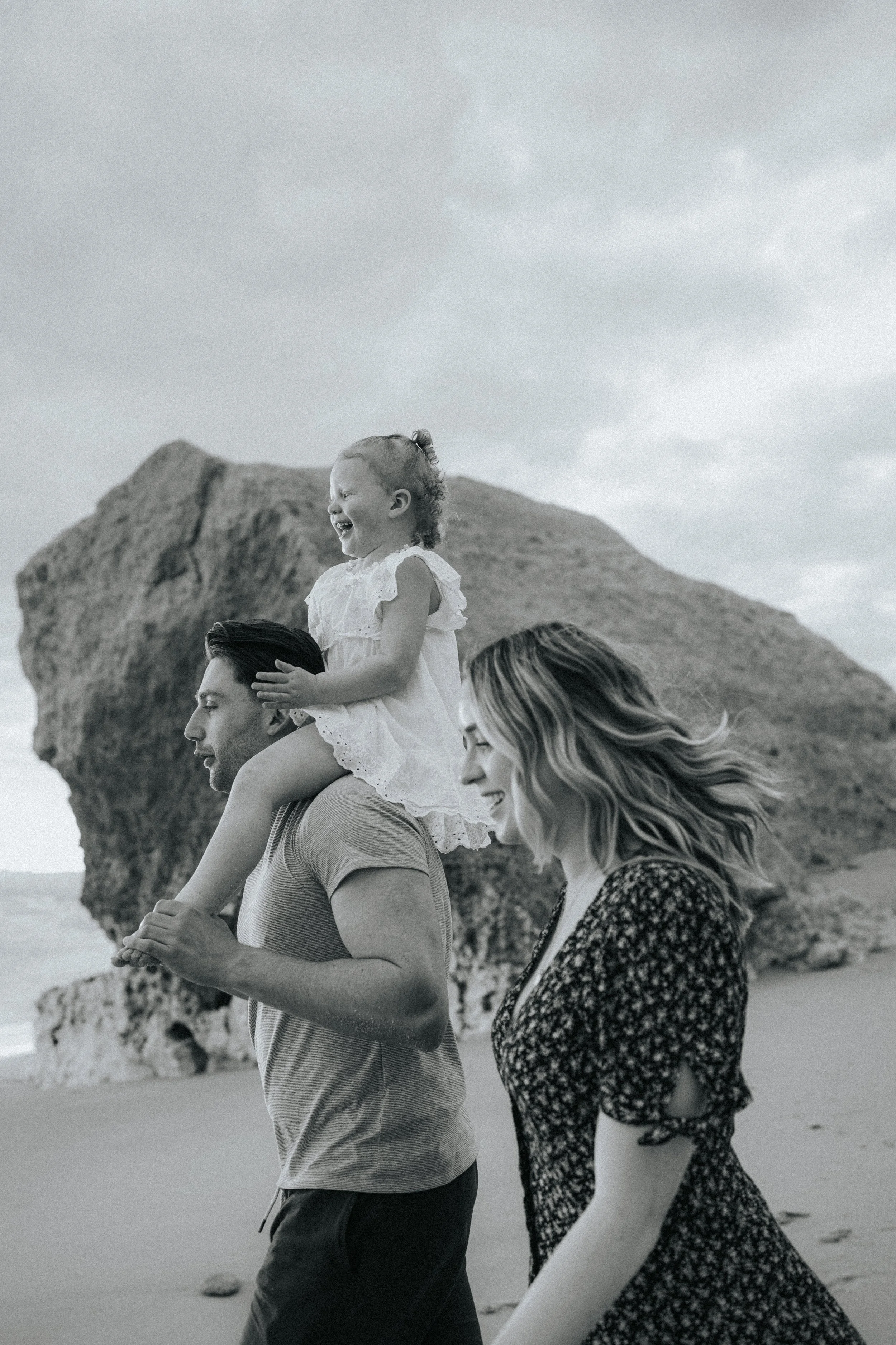 A joyful young girl with curly hair riding on the shoulders of a smiling man on a beach, with a large rock formation and cloudy sky in the background.