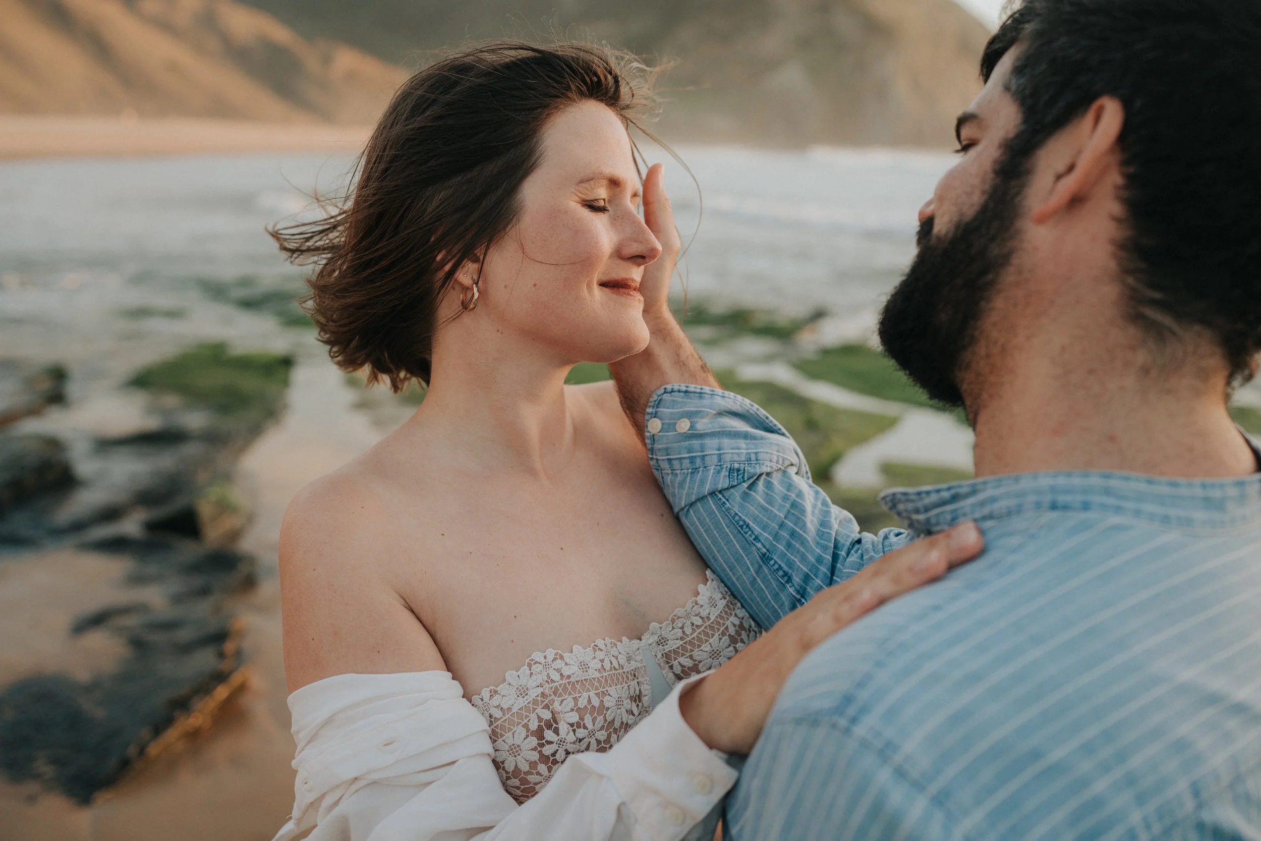 A woman with short brown hair and a man with dark hair and a beard are at the beach, with the woman smiling and eyes closed as the man's hand gently touches her face. The woman is wearing a white off-shoulder top with lace, and the man is wearing a b
