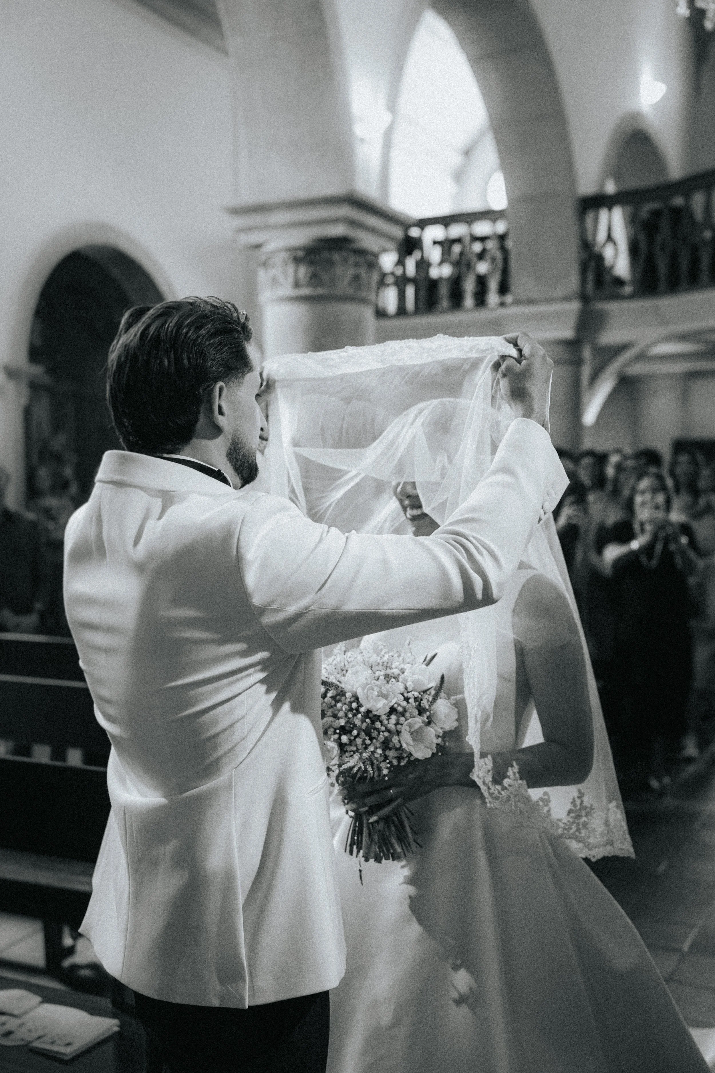 A black and white photo of a wedding ceremony with the groom lifting the bride's veil. The bride holds a bouquet of flowers, and guests are visible in the background inside a grand church with arches and ornate details.