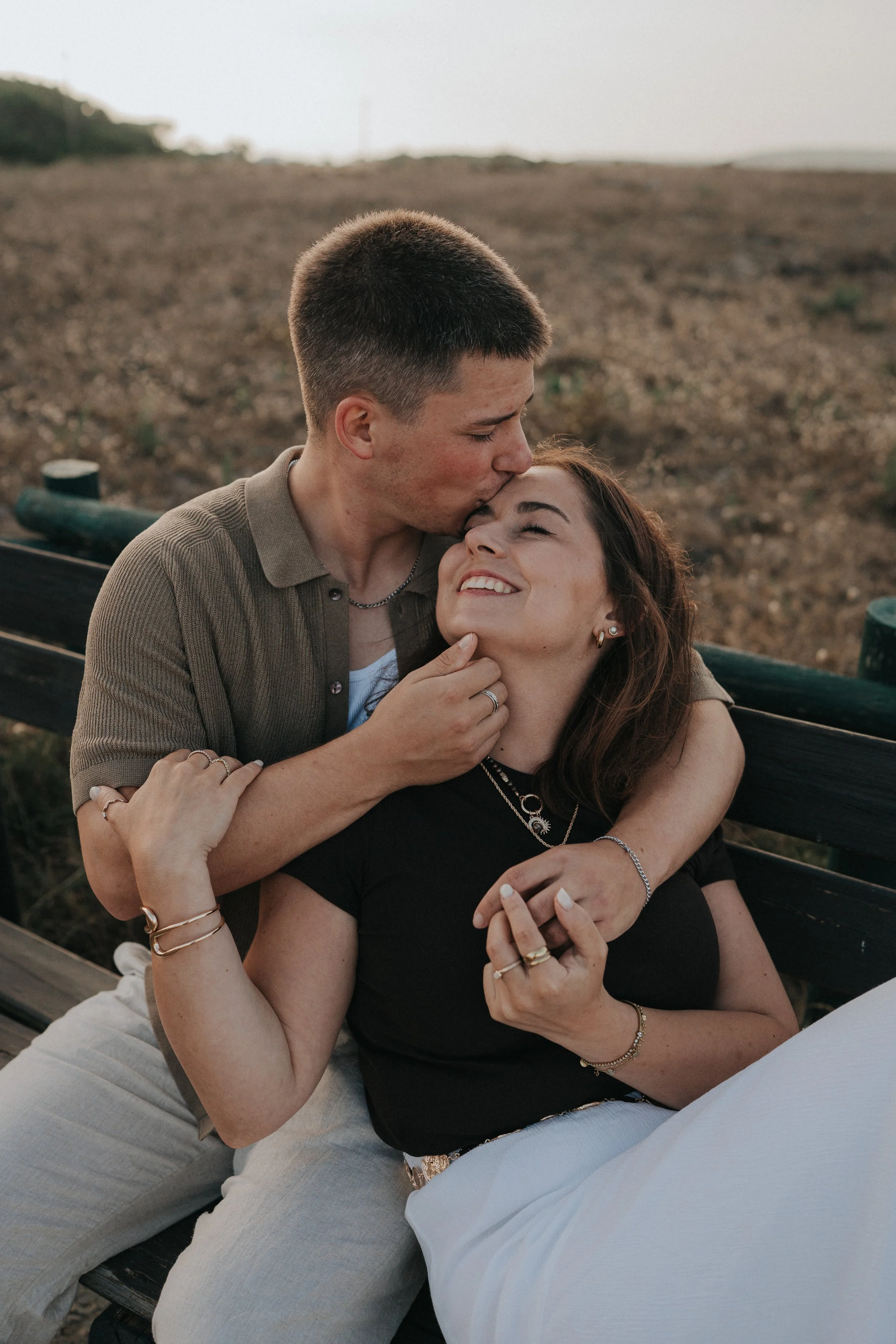 A young man and a woman sharing an affectionate moment outdoors. The man is kissing the woman on the forehead while she looks joyful and relaxed, sitting on a black bench with a natural, open landscape in the background.