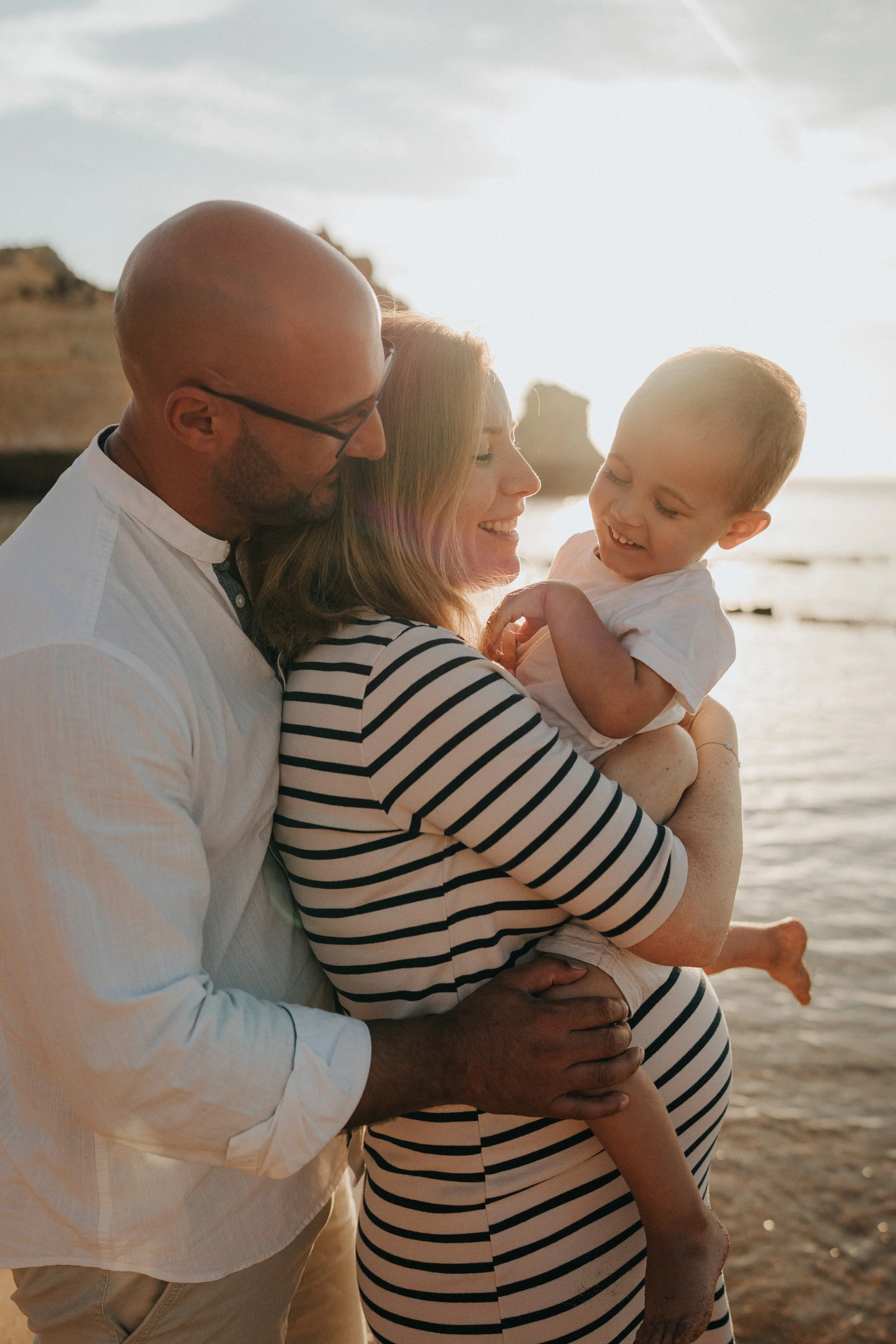 A family enjoying a moment together on the beach during sunset, with the father, mother, and young child smiling and embracing.