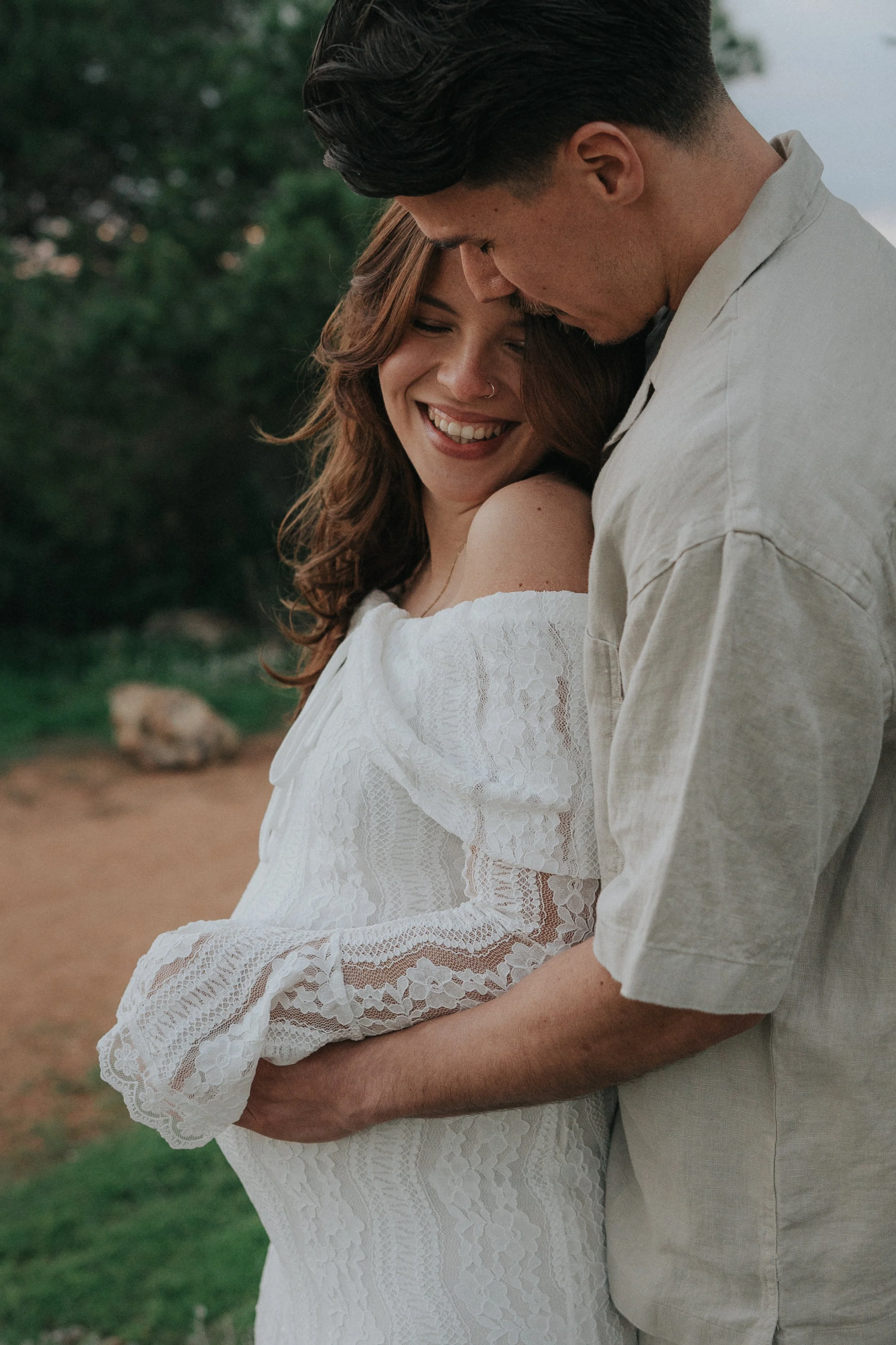 A couple, a woman with brown wavy hair and a man with dark hair, smiling and embracing outdoors. The woman wears a white lace dress with off-the-shoulder sleeves, and the man wears a light-colored shirt. They are close together, with gentle expressio