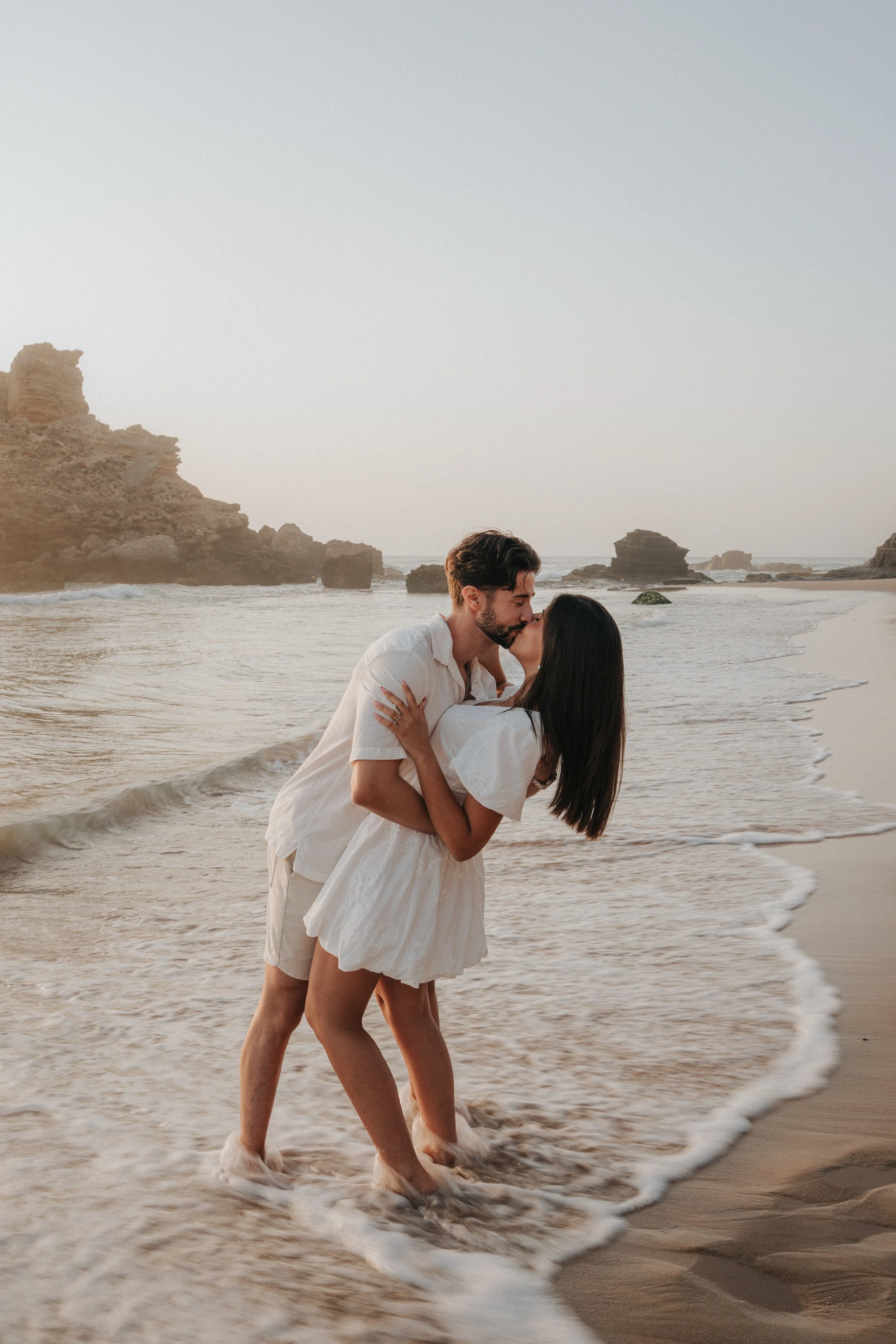 A couple kissing on the beach, walking in the shallow water, with rocky formations in the background during sunset.
