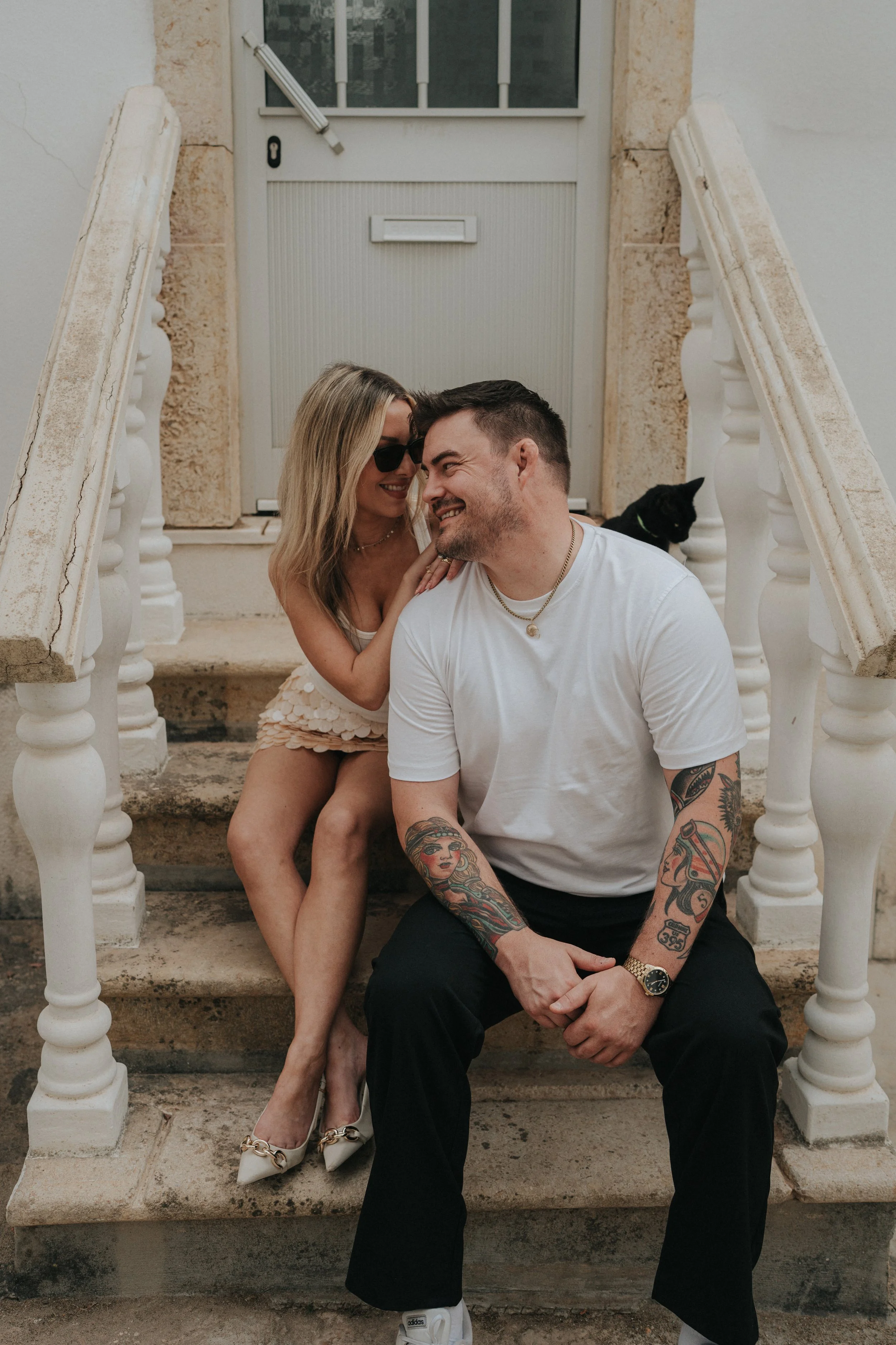 A couple sitting on front steps of a house. The woman is leaning towards the man, smiling while wearing sunglasses and a light dress. The man has tattoos on his arms, short hair, and is smiling back. A black cat sits to the right behind them.