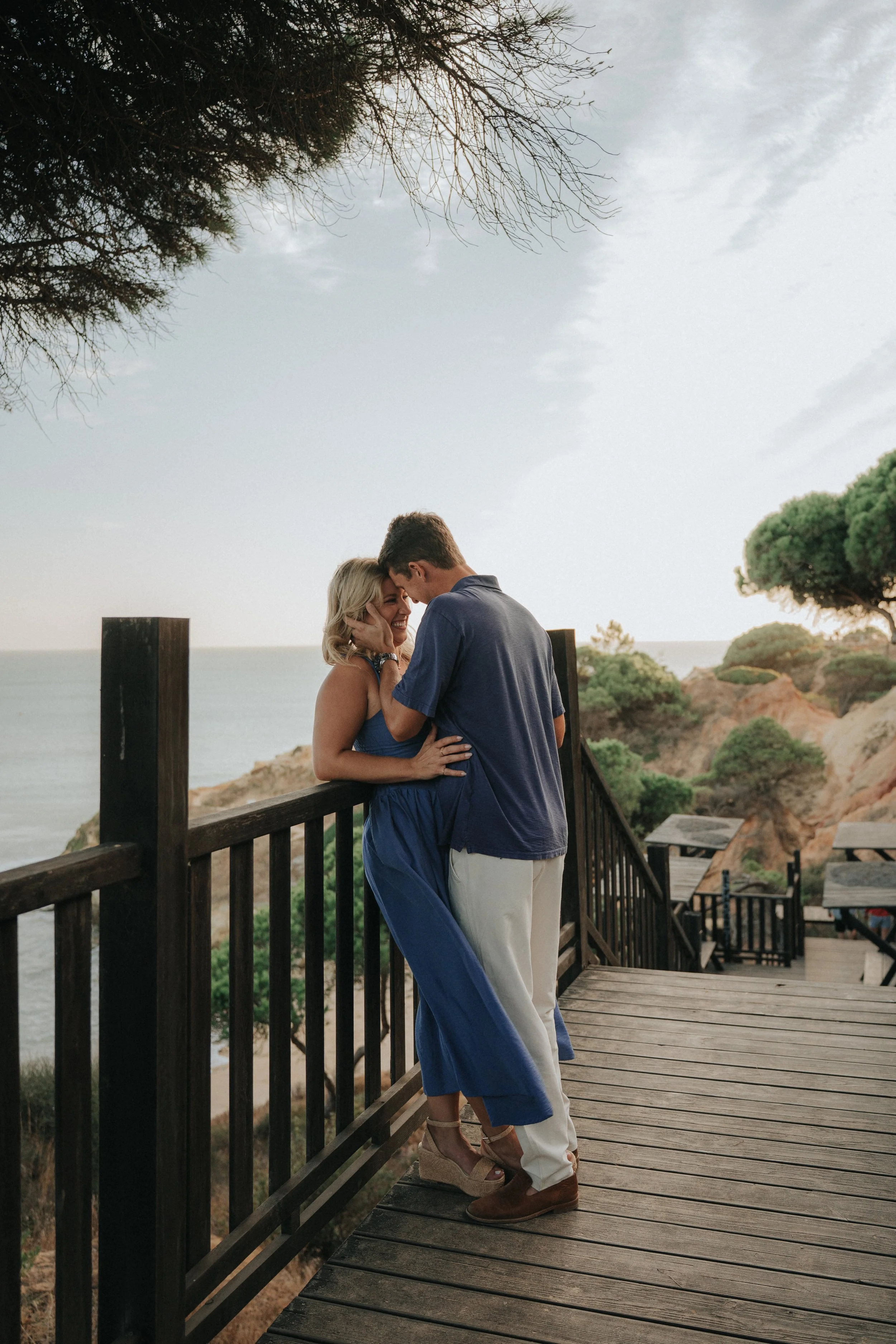 A couple embraces on a wooden boardwalk overlooking the ocean at sunset. The woman is wearing a blue dress and sandals, and the man is dressed in a dark shirt and light-colored pants. The background features trees, rocky hills, and a cloudy sky.