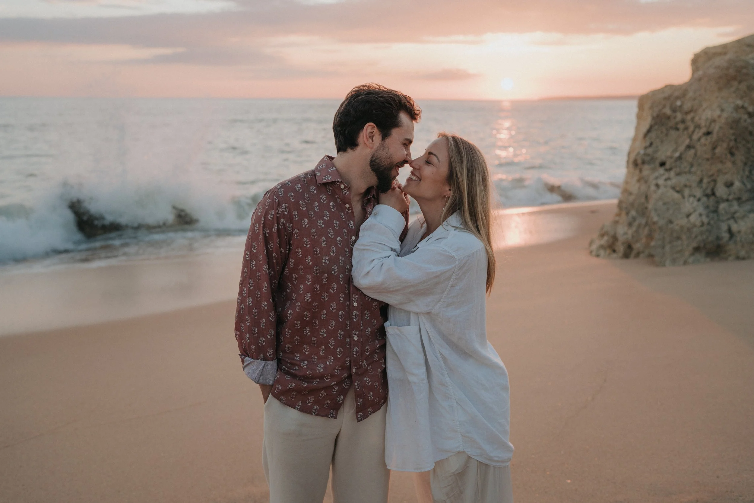 A young couple standing close together on a beach at sunset, smiling and touching noses, with ocean waves and a large rock in the background.
