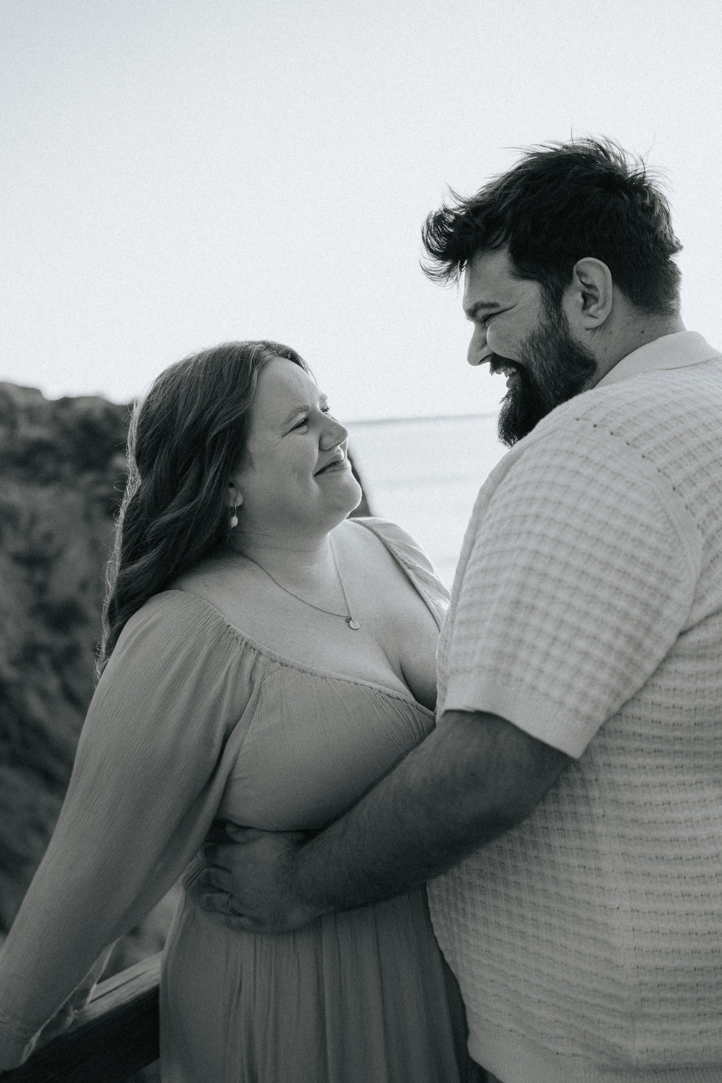 A black and white photo of a smiling couple, a woman and a man, holding each other close outdoors near the coast with a rocky background.