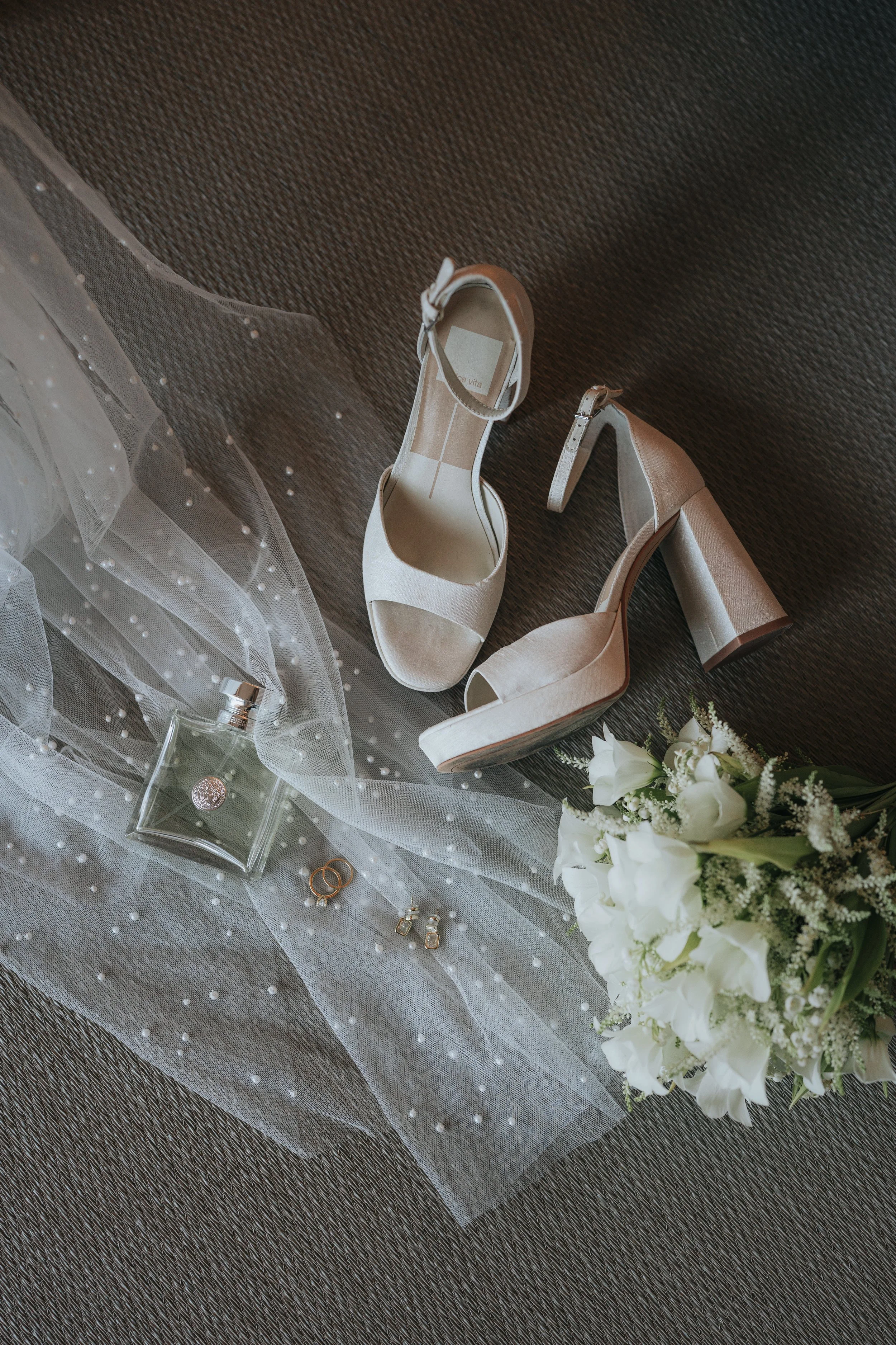 A pair of beige high-heeled shoes, a bouquet of white flowers, a small glass bottle, rings, and pearl-decorated tulle fabric on a dark textured surface, likely part of wedding accessories.