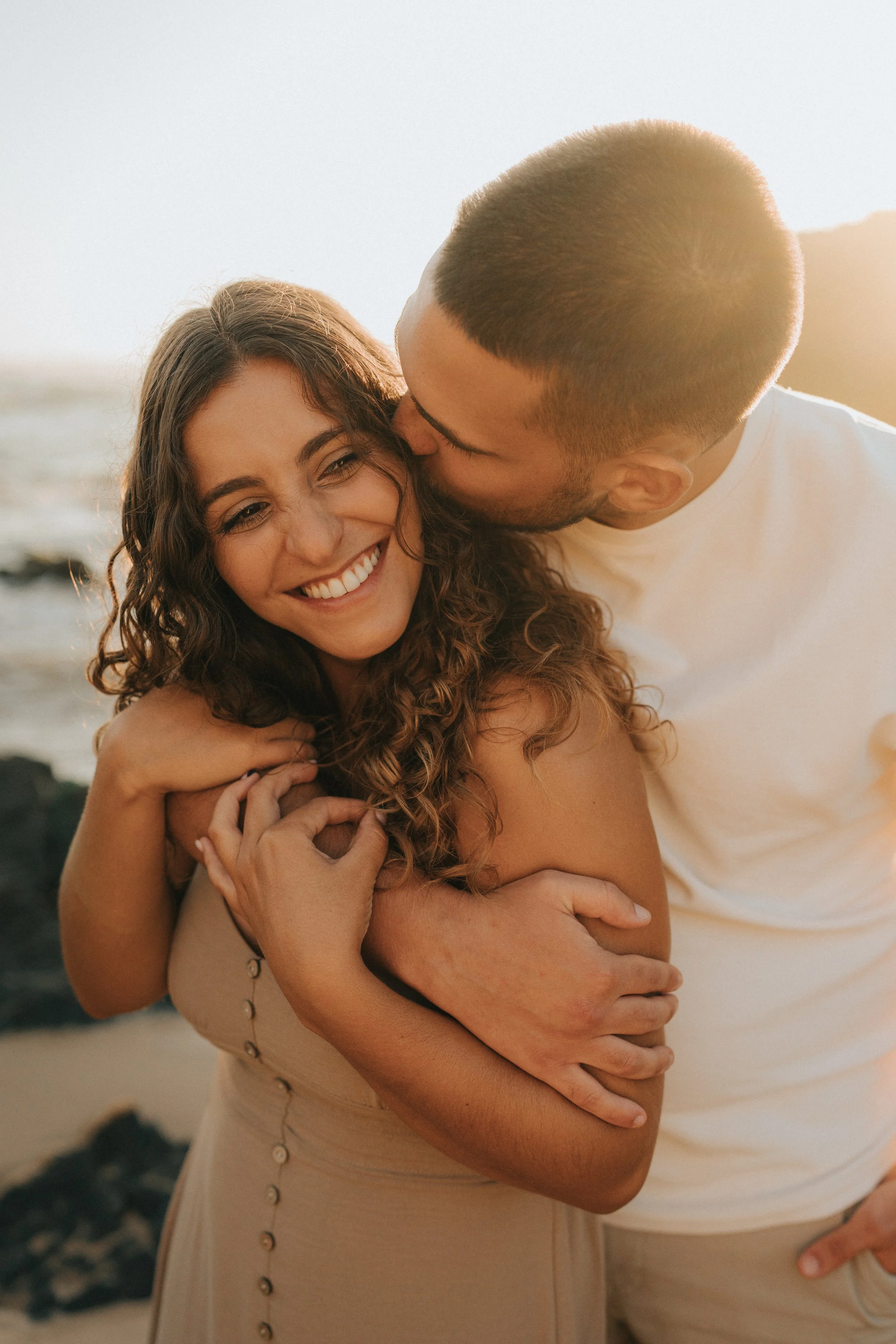 A couple embracing at the beach during sunset, with the man kissing the woman on her cheek and both smiling.
