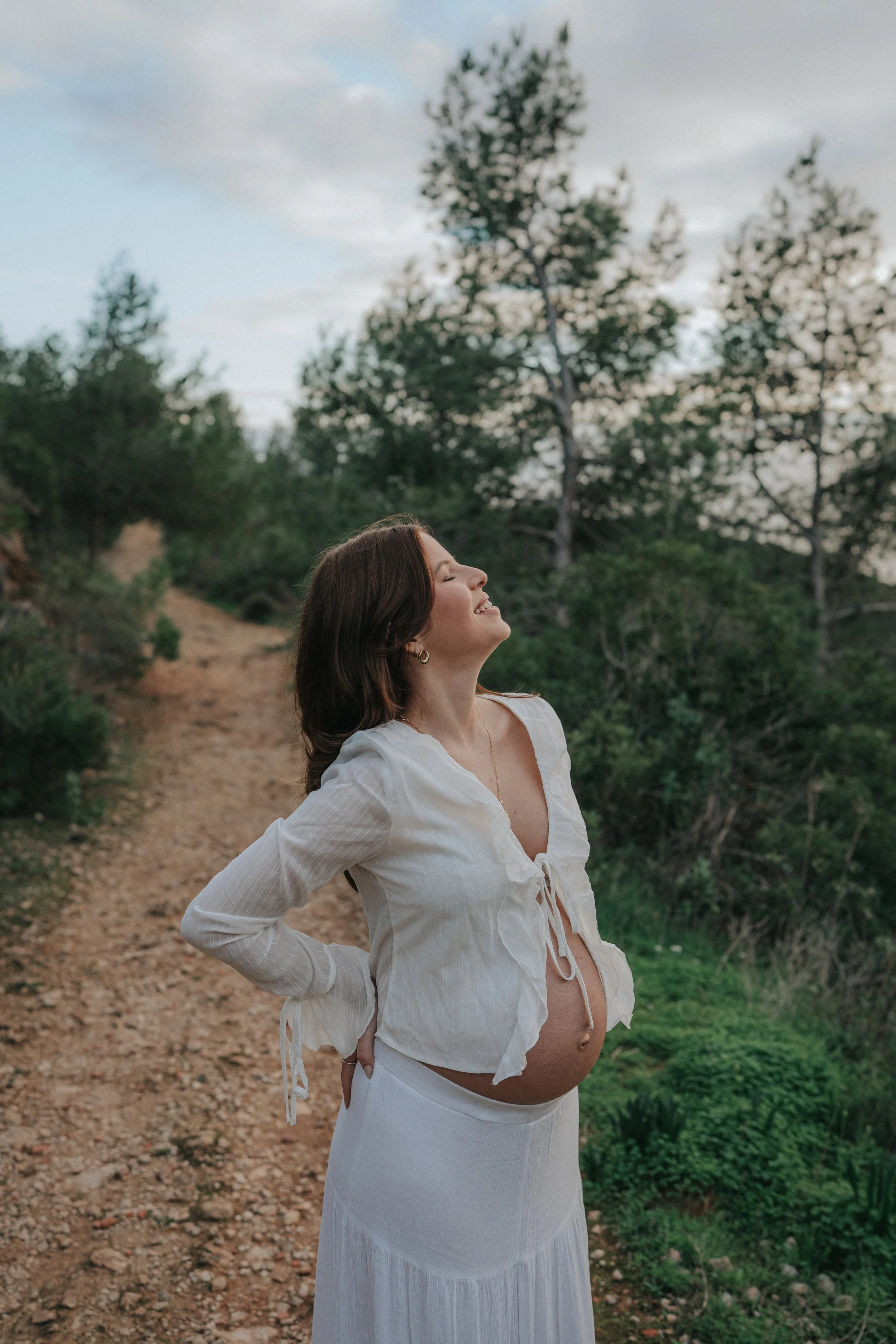 A pregnant woman with brown hair smiling and looking upwards, standing outdoors on a dirt path surrounded by trees and greenery, wearing a white open-collared blouse and a long white skirt.