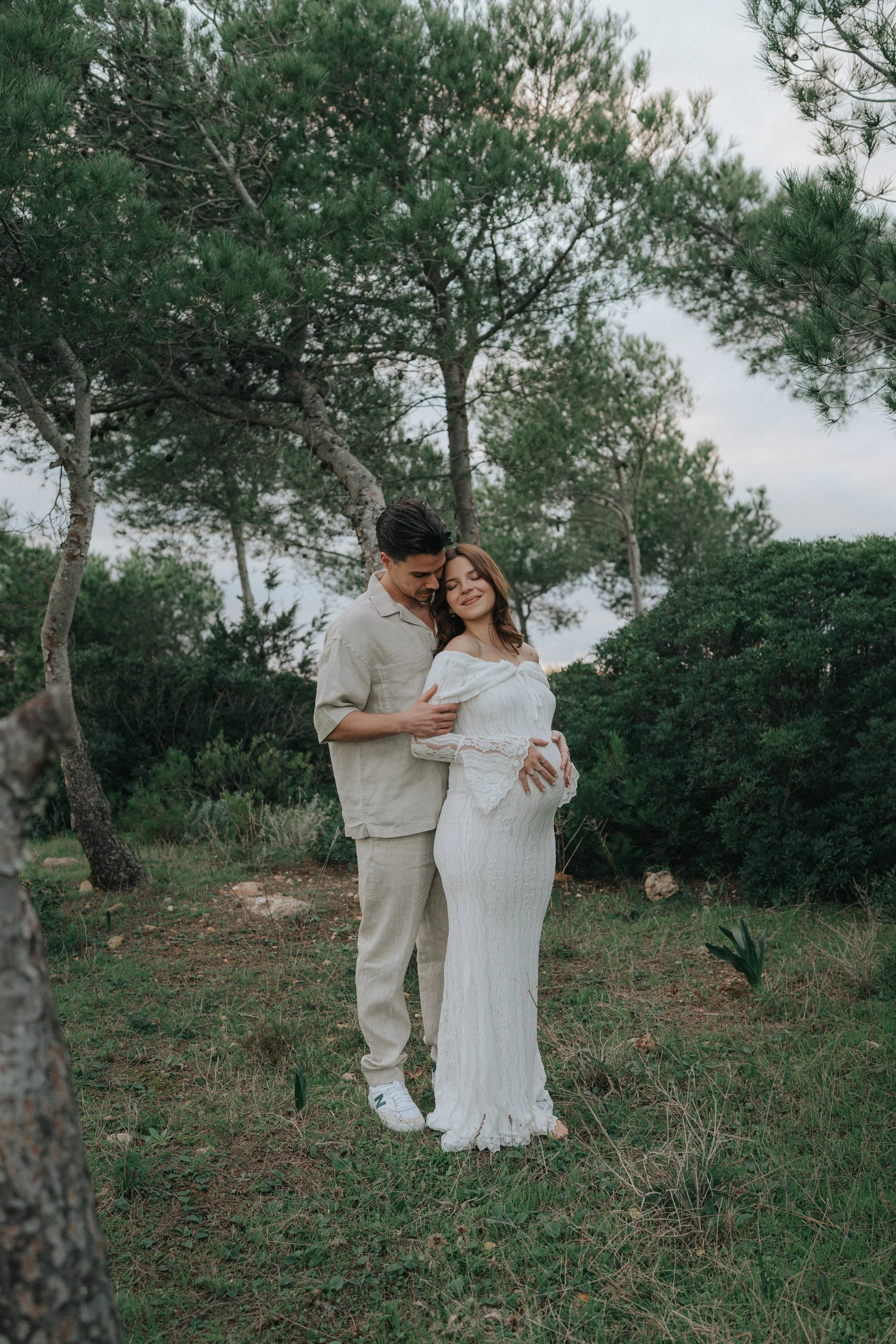 A couple stands close together outdoors under a canopy of trees, smiling and embracing, with the woman holding her pregnant belly.