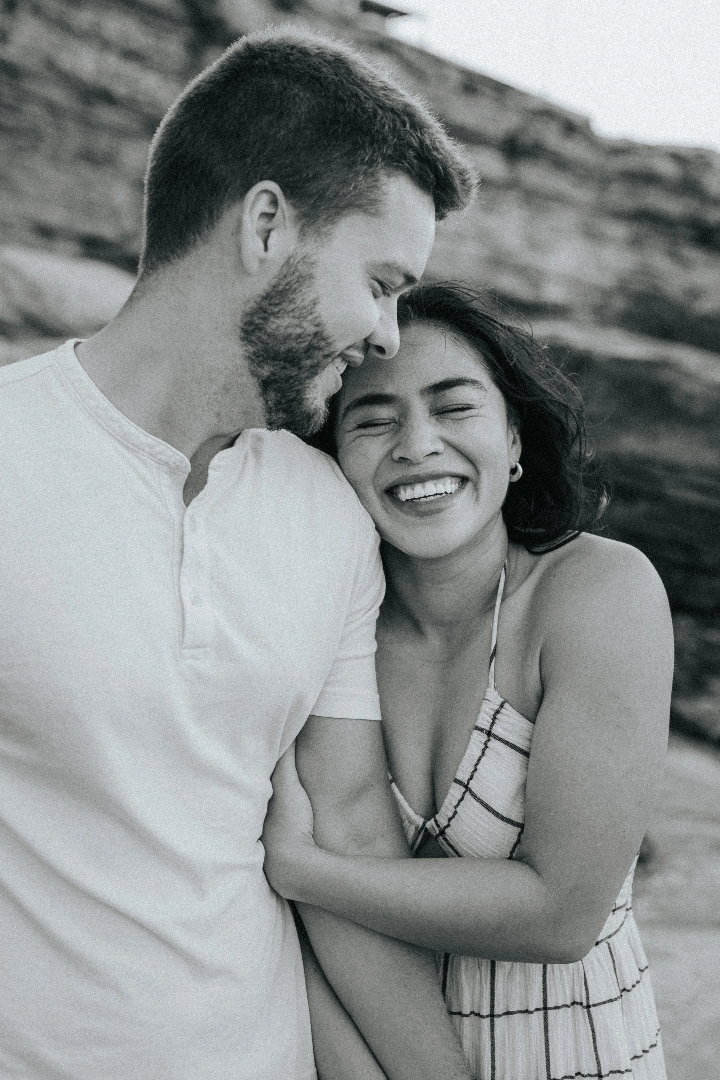 A black and white photo of a joyful couple smiling and hugging outdoors, with a rocky background.