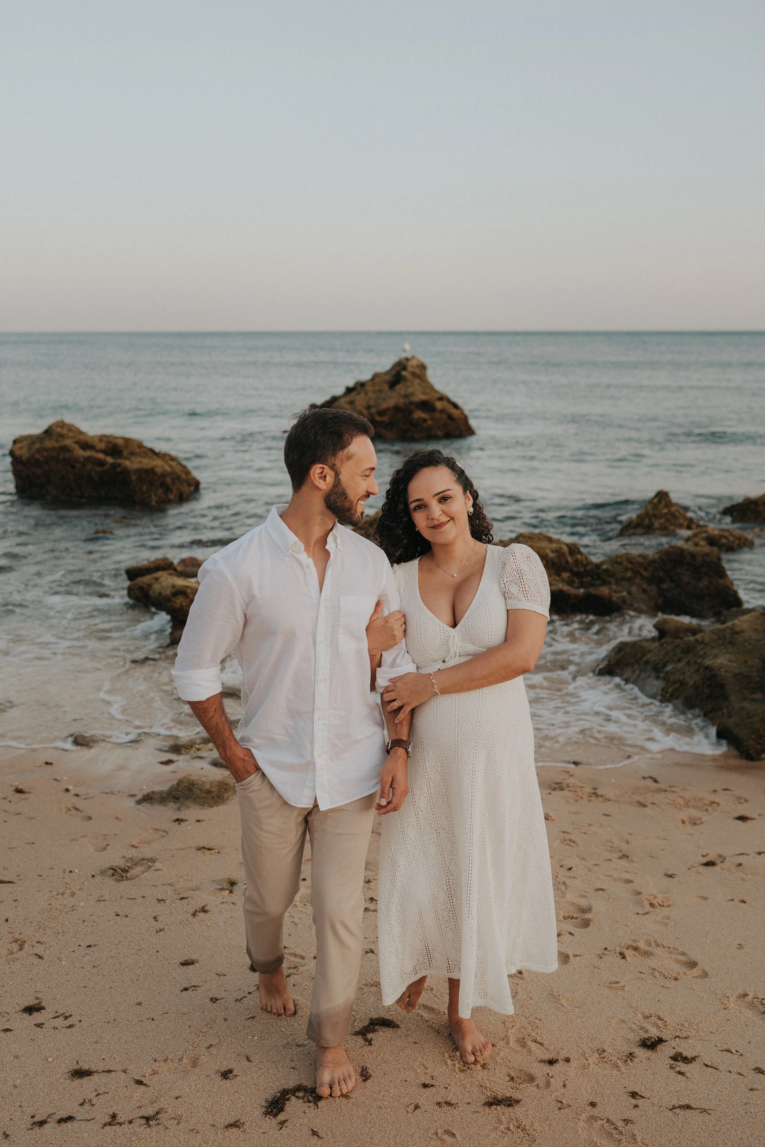 A couple standing barefoot on the sandy beach near rocks with the ocean in the background, smiling and enjoying a scenic sunset or sunrise.
