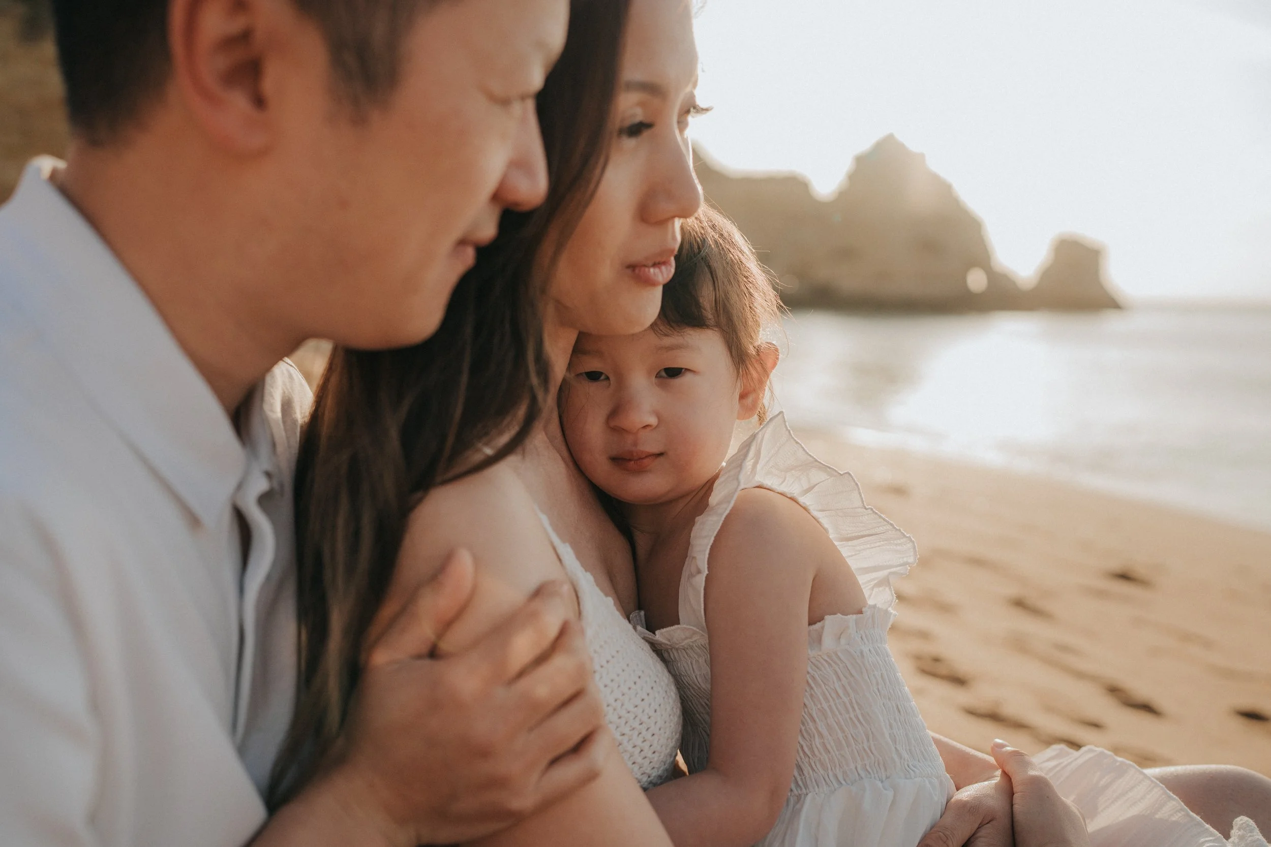 A family of three sitting on the beach, close together, looking out at the ocean and rocky formations in the distance, during sunset.