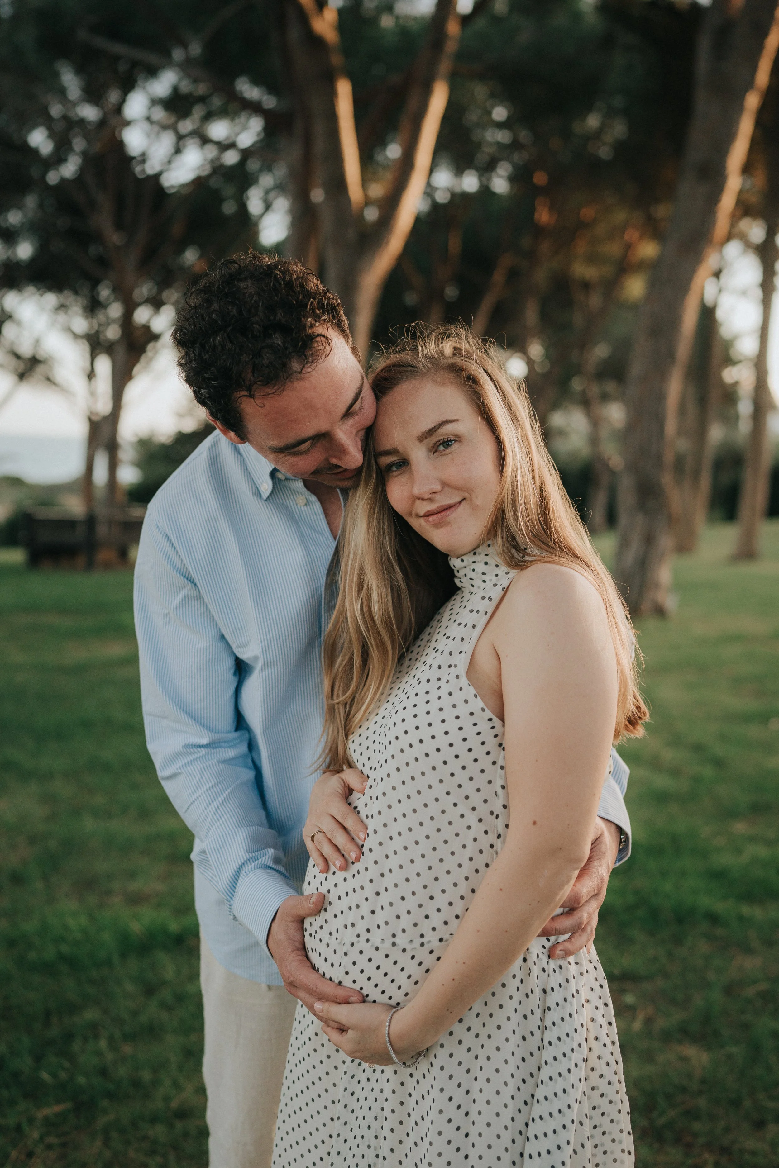 A young pregnant woman with long blonde hair, wearing a white polka dot dress, standing outdoors with two men, one embracing her from behind and the other touching her belly, under a canopy of trees during daylight.