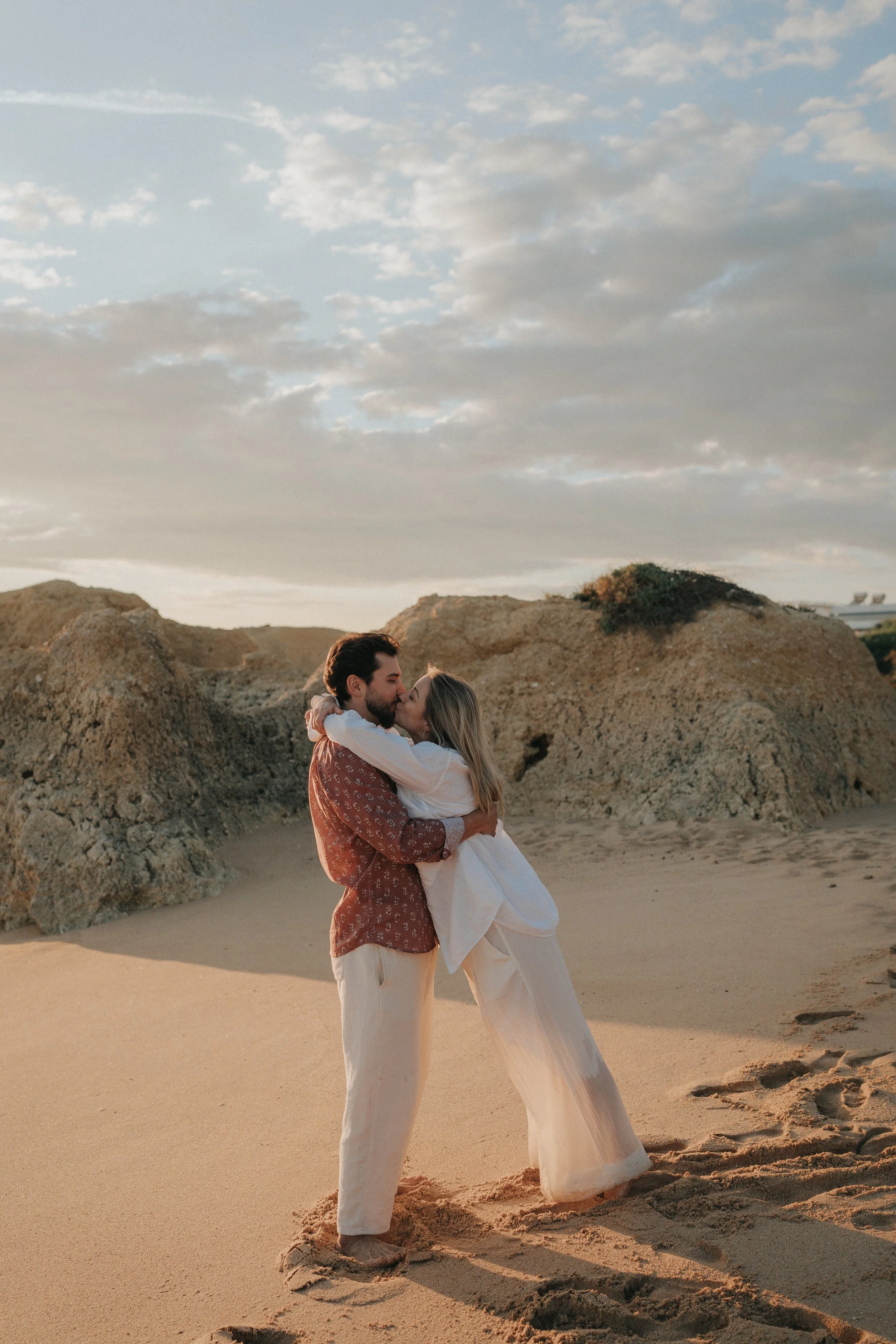 A couple embracing and sharing a kiss on a sandy beach with large rocks, under a partly cloudy sky during sunset or sunrise.