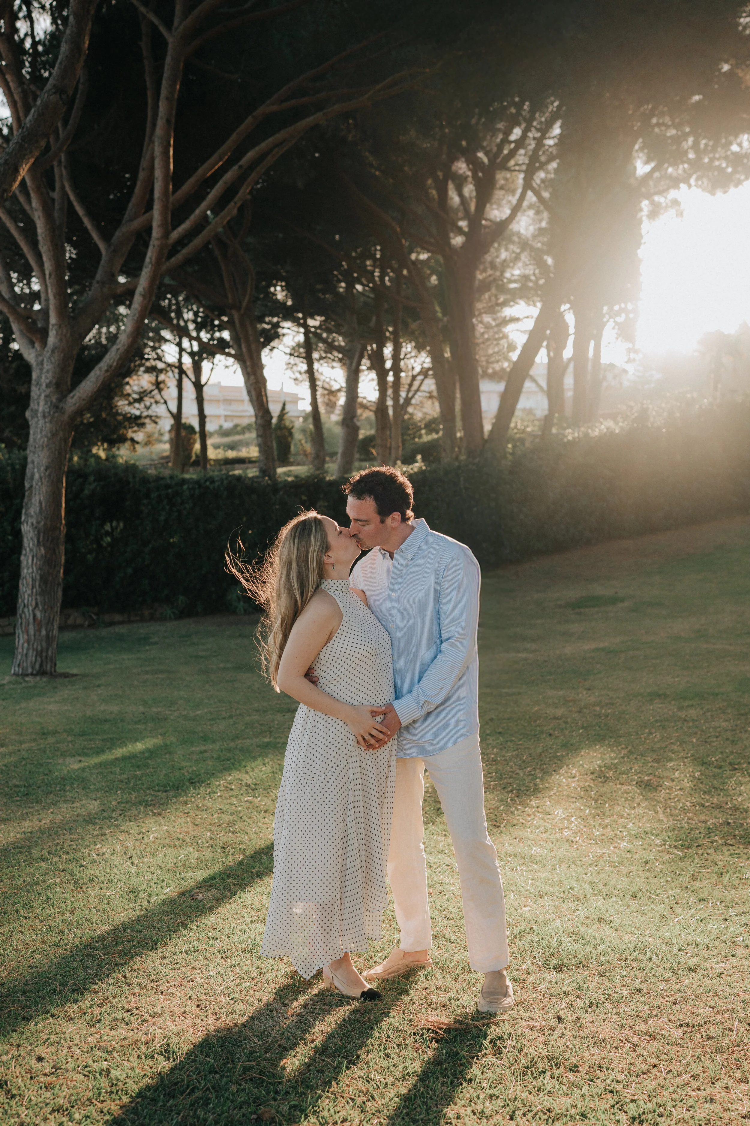A couple stands closely together outdoors during sunset, kissing. The woman is wearing a white polka dot dress and heels, and the man is in light-colored pants and a blue shirt. Tall trees and a grassy area are in the background.