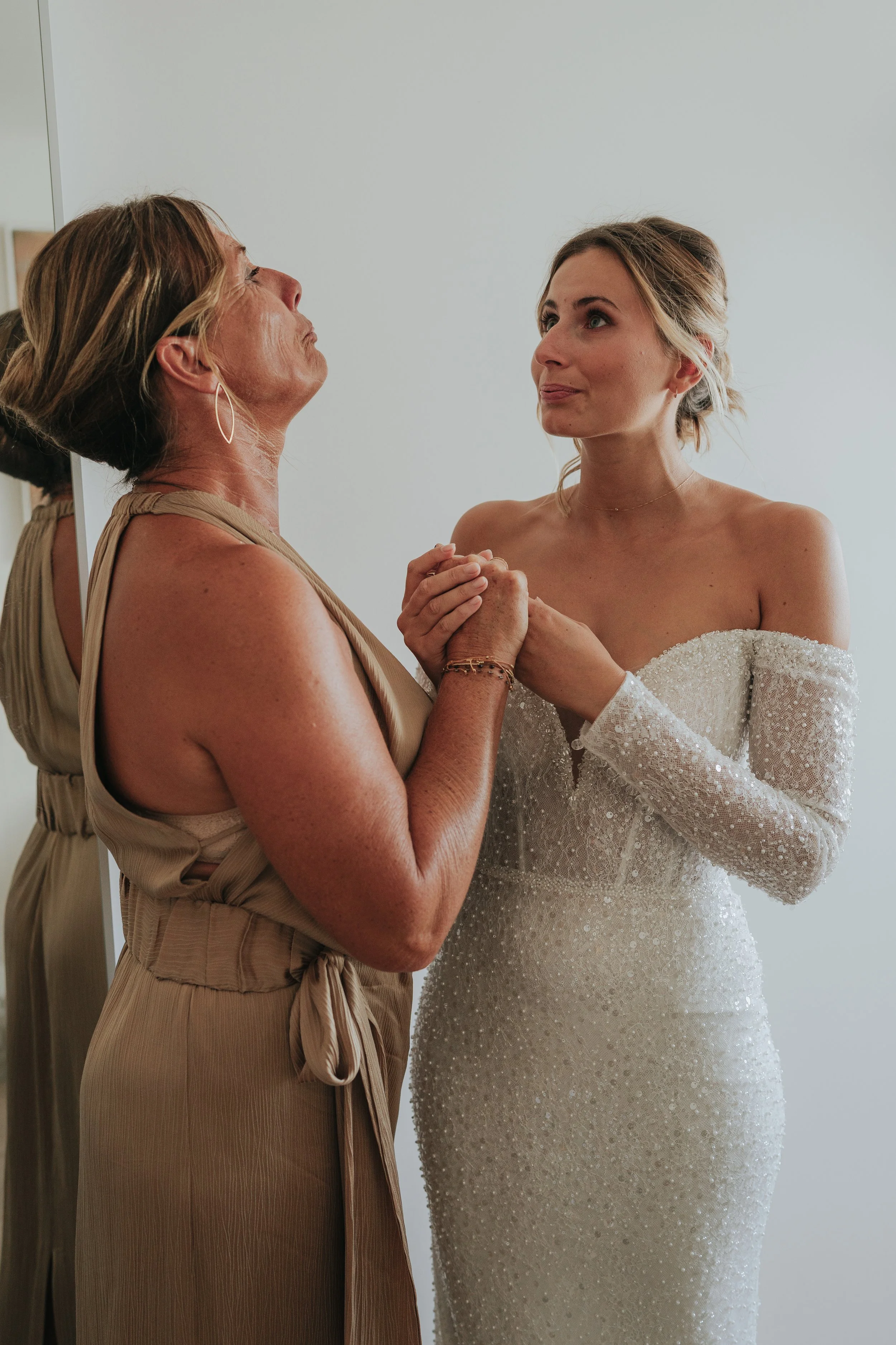 A woman in a beige dress and hoop earrings is holding hands with a young woman in a white sparkly dress, possibly at a wedding or special event, against a plain gray background.