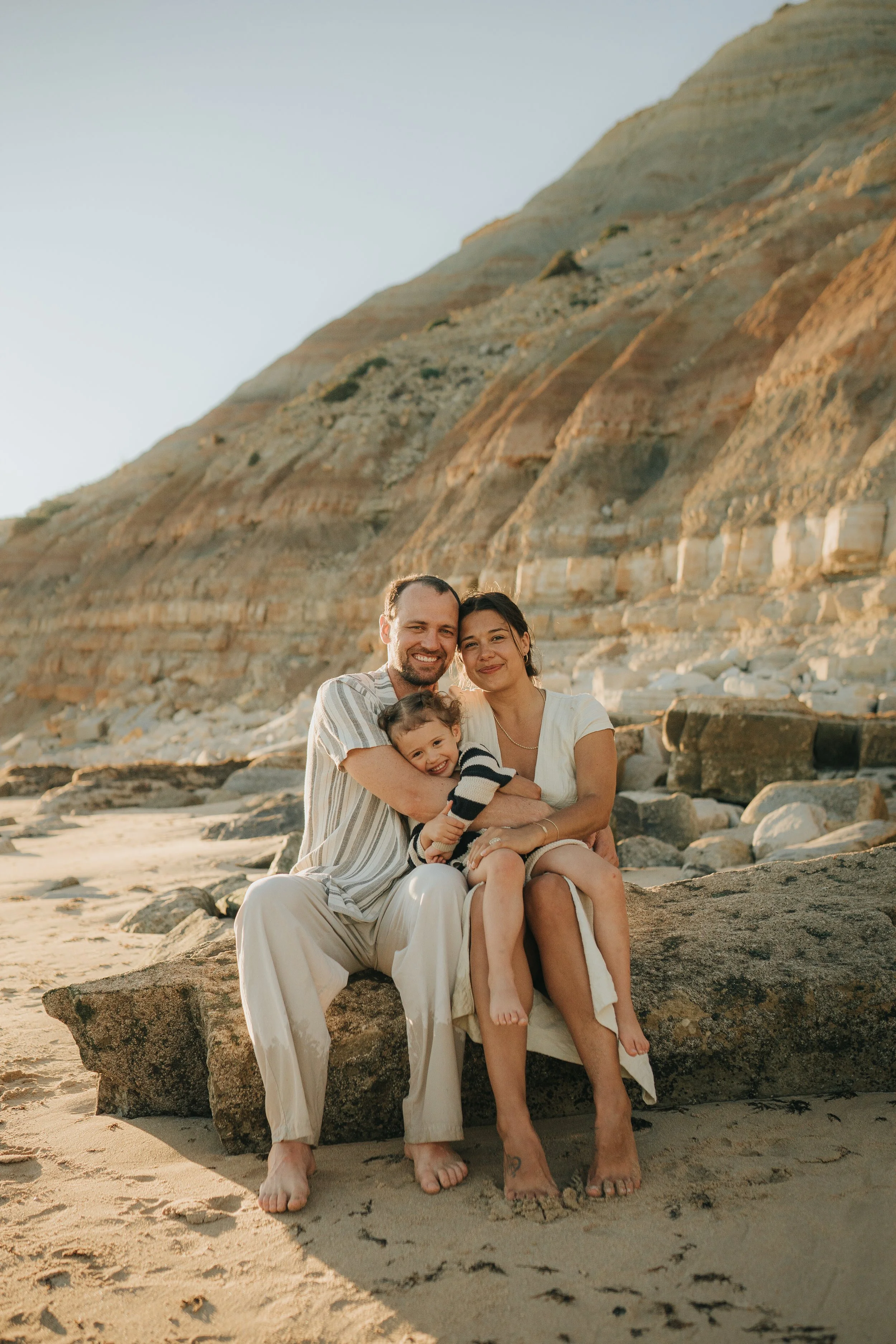 A family of three sitting on a large rock at the beach, with a cliff in the background under a clear sky. The man and woman are smiling, hugging their young child.