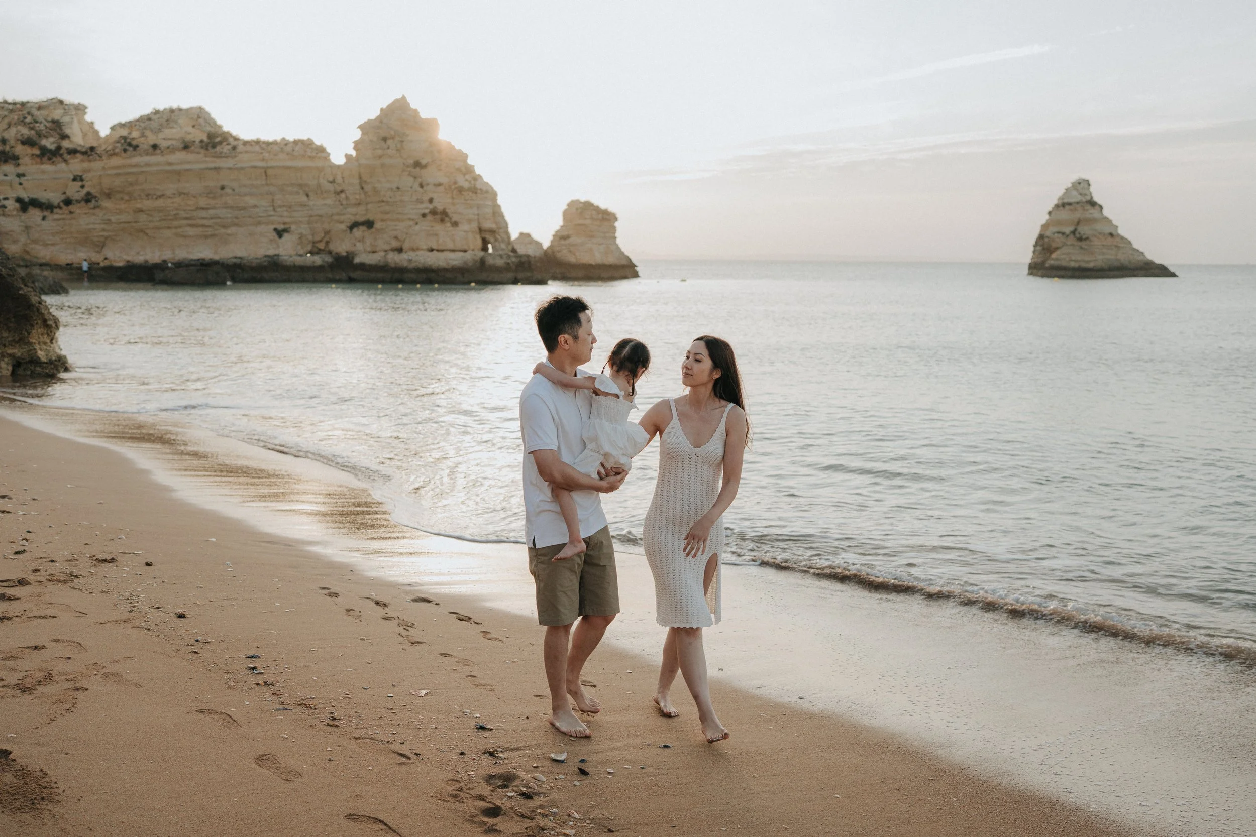 A family of three walking on the beach during sunset, with rocky cliffs in the background and the ocean.