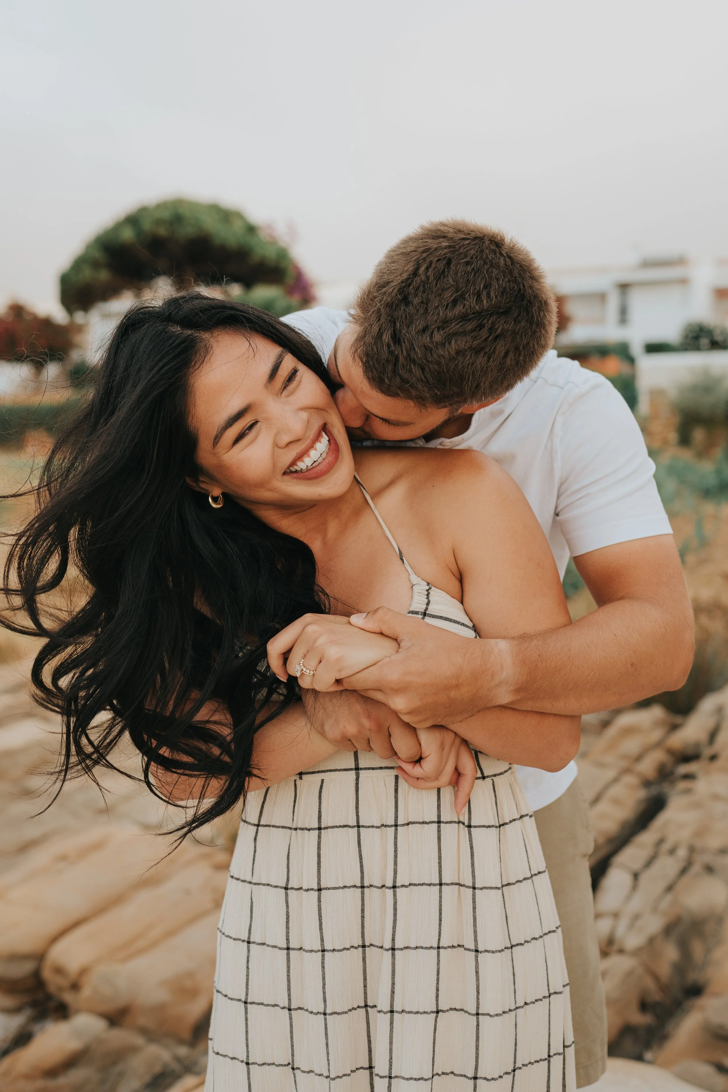 A happy couple hugging outdoors with rocks and a tree in the background.